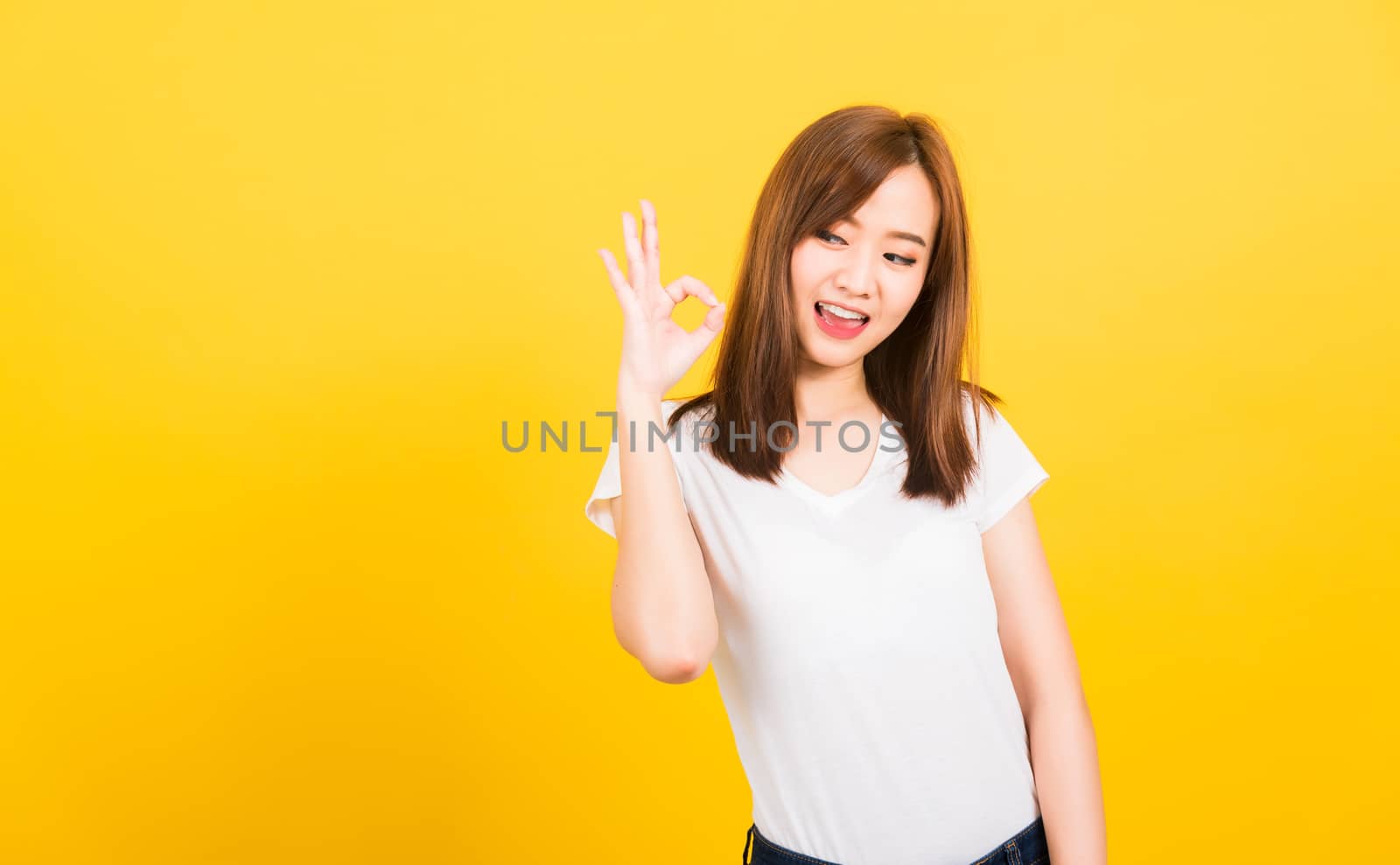 Asian happy portrait beautiful cute young woman teen standing wear t-shirt showing gesturing ok sign with fingers looking to side isolated, studio shot on yellow background with copy space for text