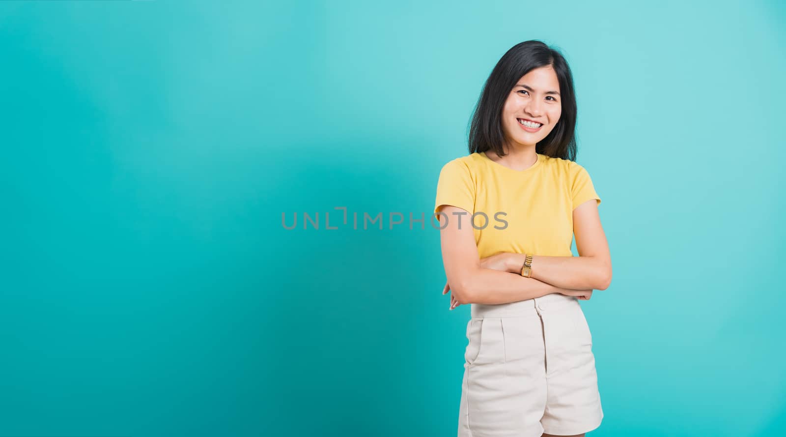 Portrait Asian beautiful young woman standing smile seeing white teeth, She crossed her arms and looking at camera, shoot photo in studio on blue background. copy space to put text on left-hand side.