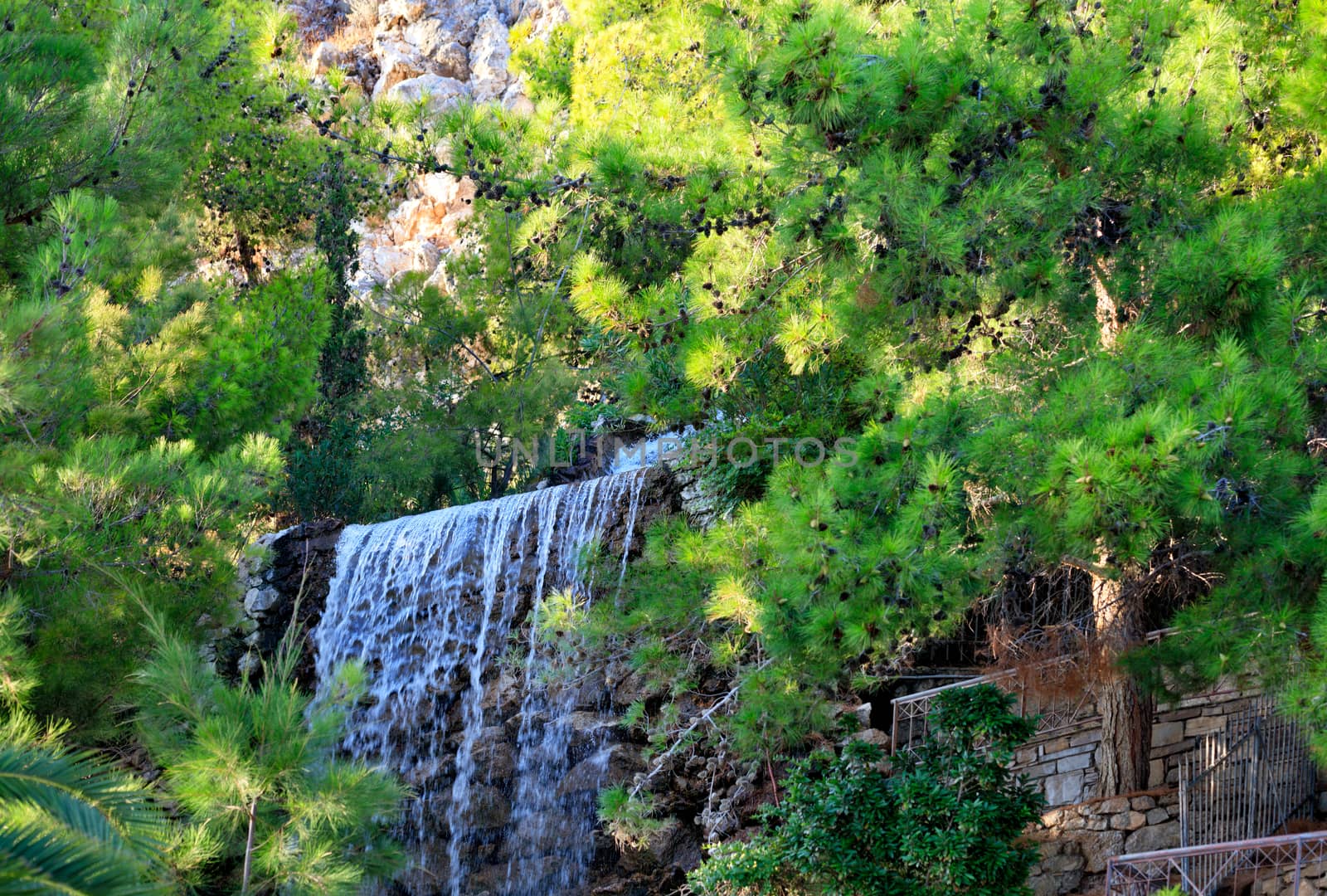 A large waterfall with radon water among boulders and thickets of spruce at the foot of a mountain in Loutraki, Greece. by Sergii