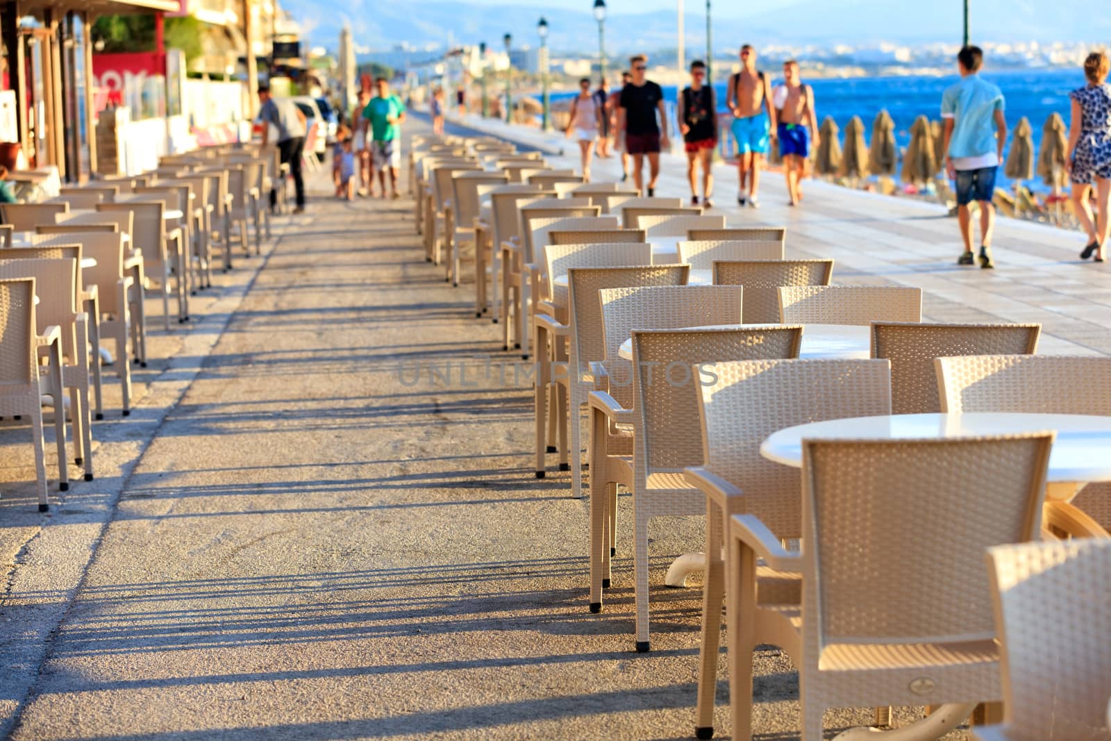 Tables and chairs of the beach cafe are located along the seafront, along which resting people and children walk in blur, image with copy space.