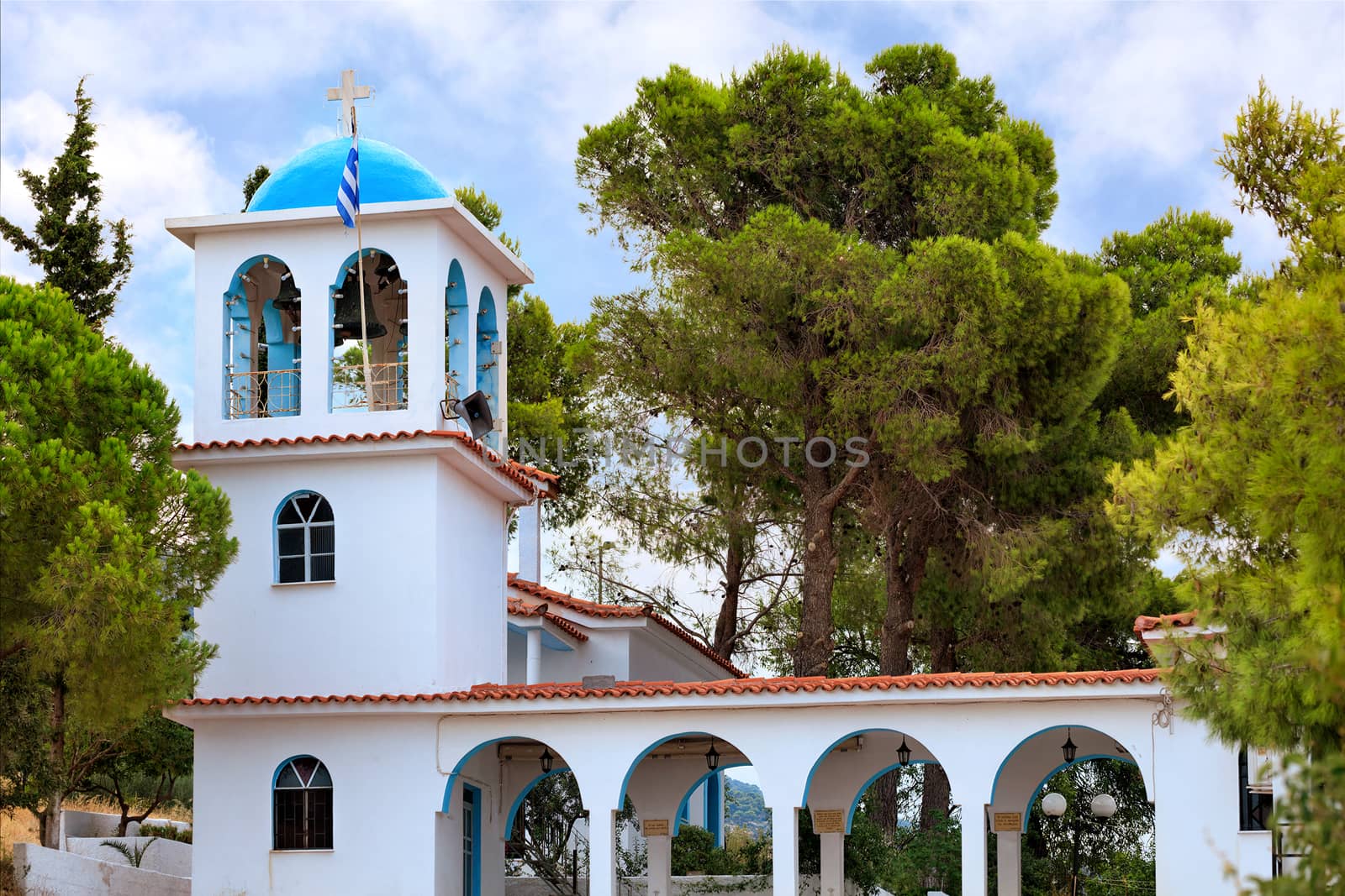 The ritual bell tower of the Orthodox Church of Ekklisia Agios Ioannis in Loutraki surrounded by green Mediterranean spruces, August 15, 2019, Greece.