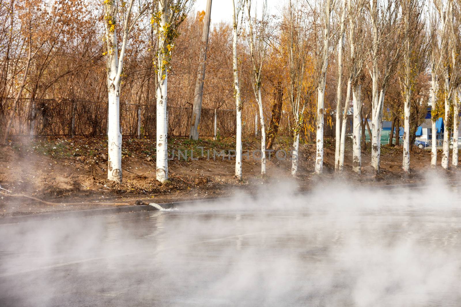 Hot water is supplied to the road using a pump and a fire hose, hot water is poured onto the road, water vapor rises tightly on the asphalt, autumn trees look at what is happening in disbelief, image with copy space.