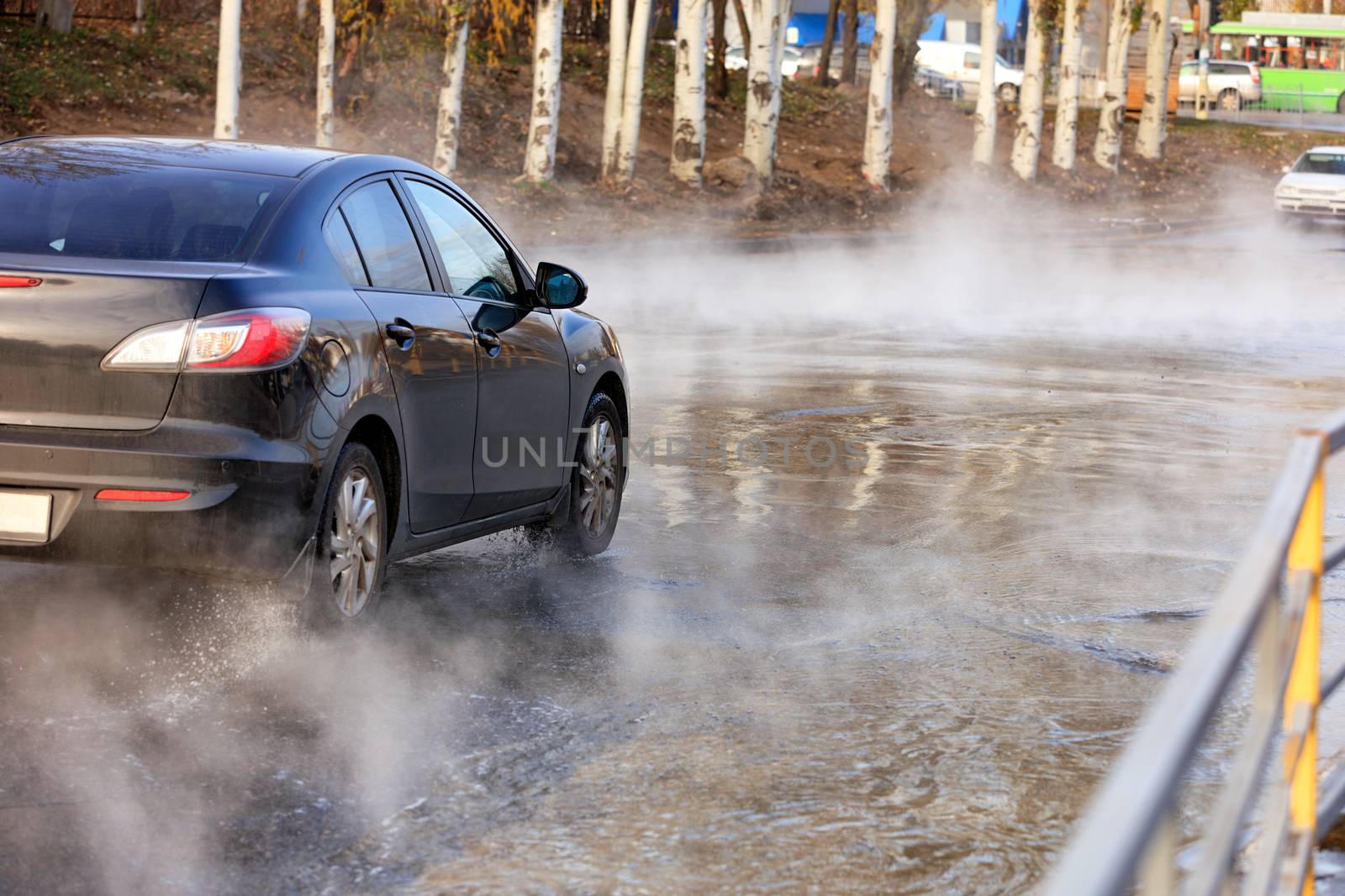 Accident on a heating main, a break in a pipe with hot water, a black car driving along a flooded road, water vapor forms a smokescreen over the asphalt, an image with copy space.