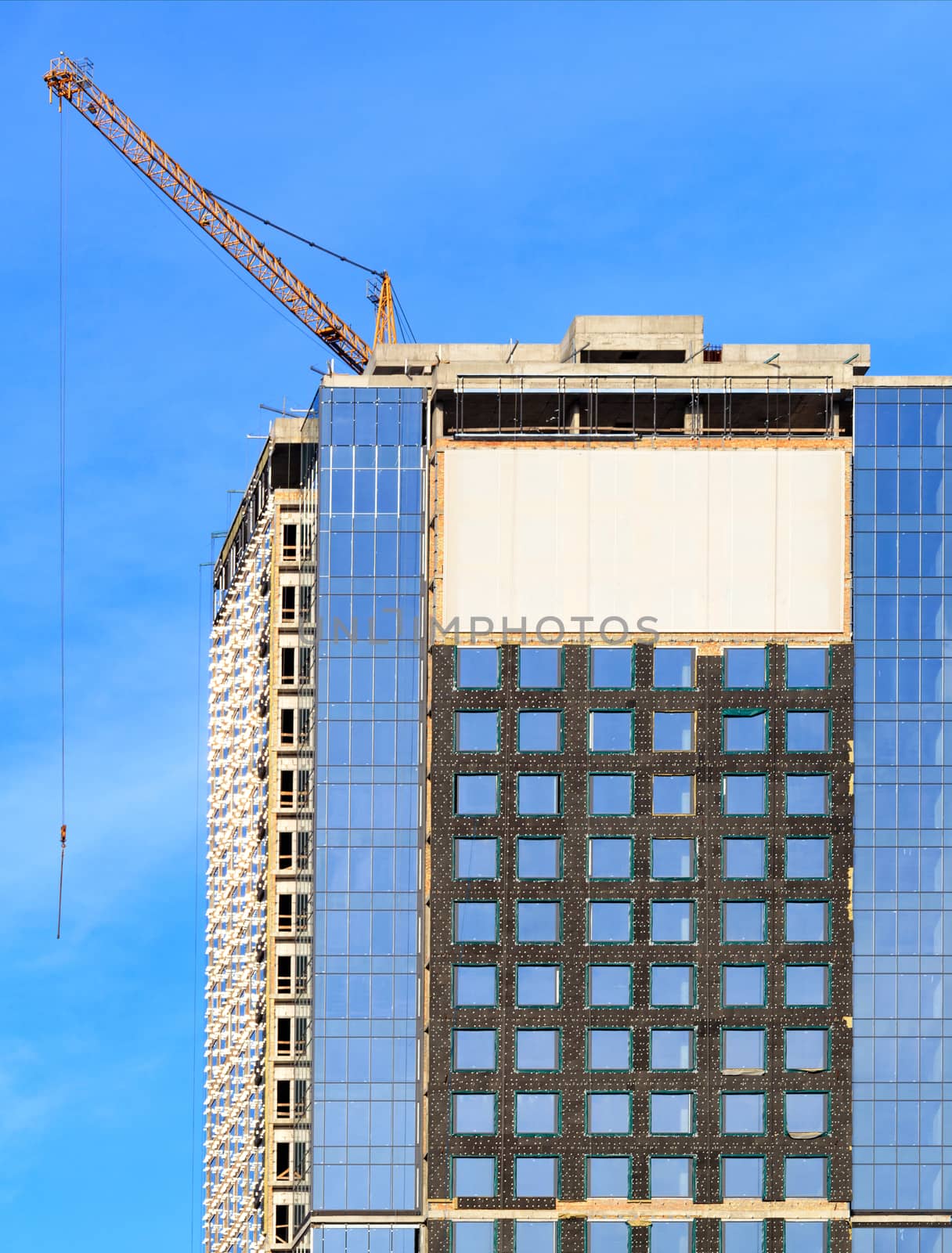 In the construction of modern residential concrete buildings with a glass facade, a tower crane is used, the blue sky is reflected in the windows of the building, vertical image with copy space.