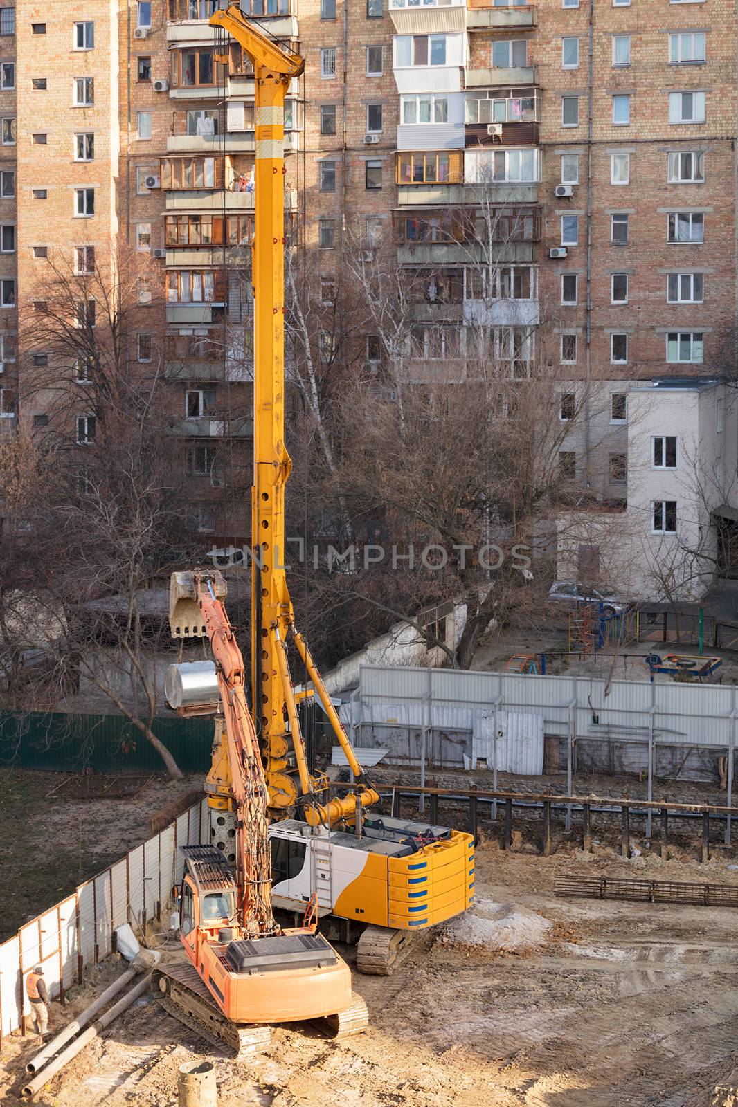 Heavy construction equipment, an excavator and a high powerful crane drill holes in the ground for laying concrete piles in the foundation of the future home, a vertical image with copy space.