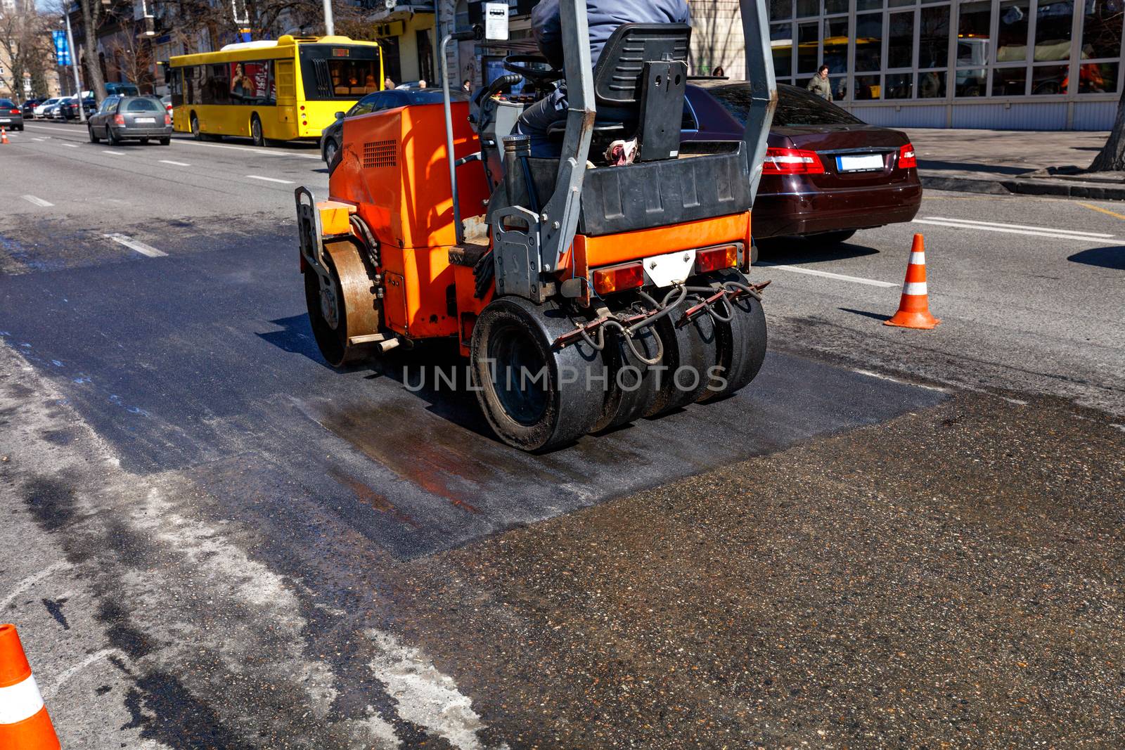 A heavy orange vibratory roller compacts and levels a section of road on a city street fenced with traffic cones, image with copy space.