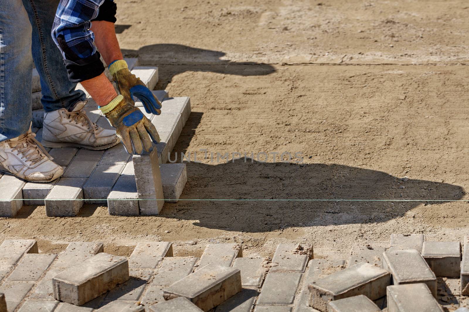 A worker lays paving slabs on a bright sunny day along a stretched cord onto prepared smooth sandy soil on the pavement, image with copy space.