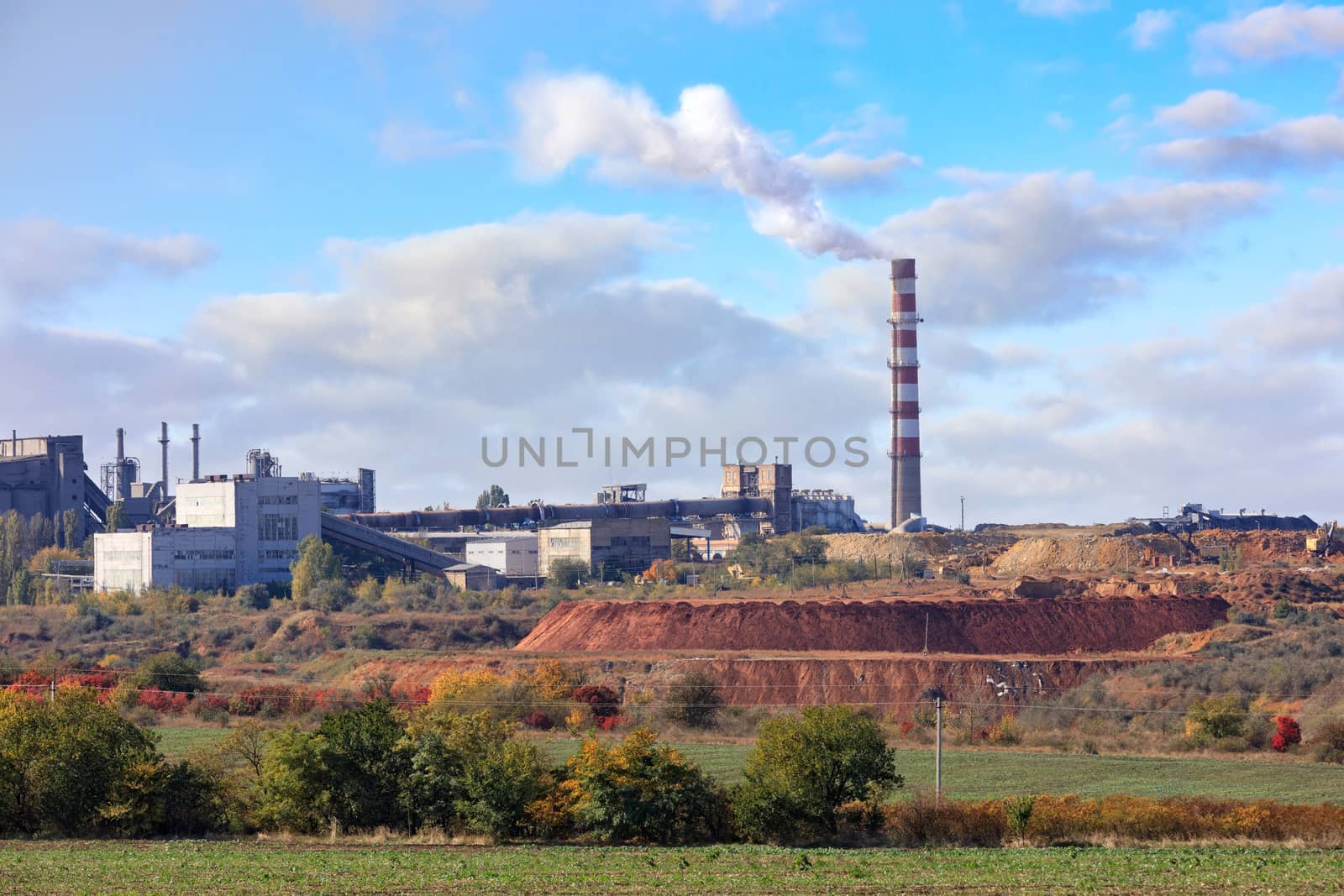 The pipe of the cement plant rises above the quarry of clay and limestone against the background of a slightly cloudy blue summer sky.