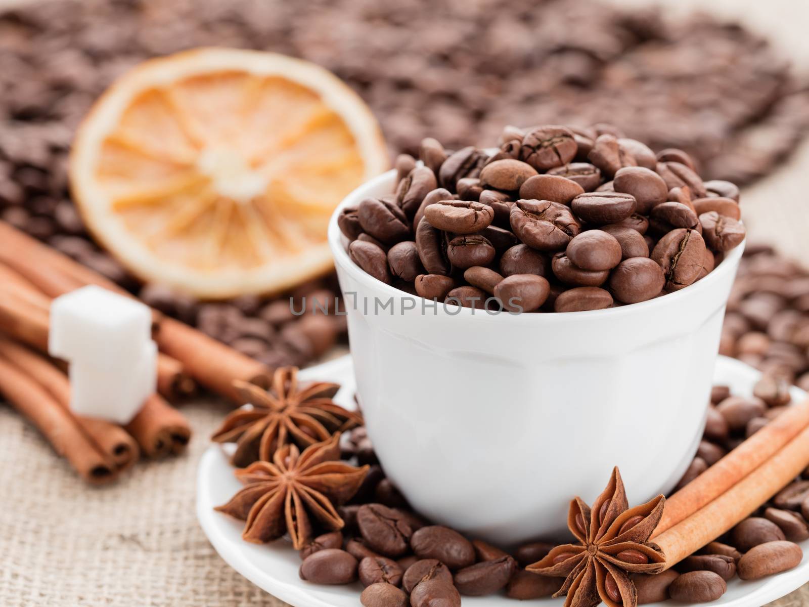 Grains of black coffee in a white porcelain dish. Cinnamon sticks and star anise on a white porcelain dish. Dried lemon slices and sugar in the background.