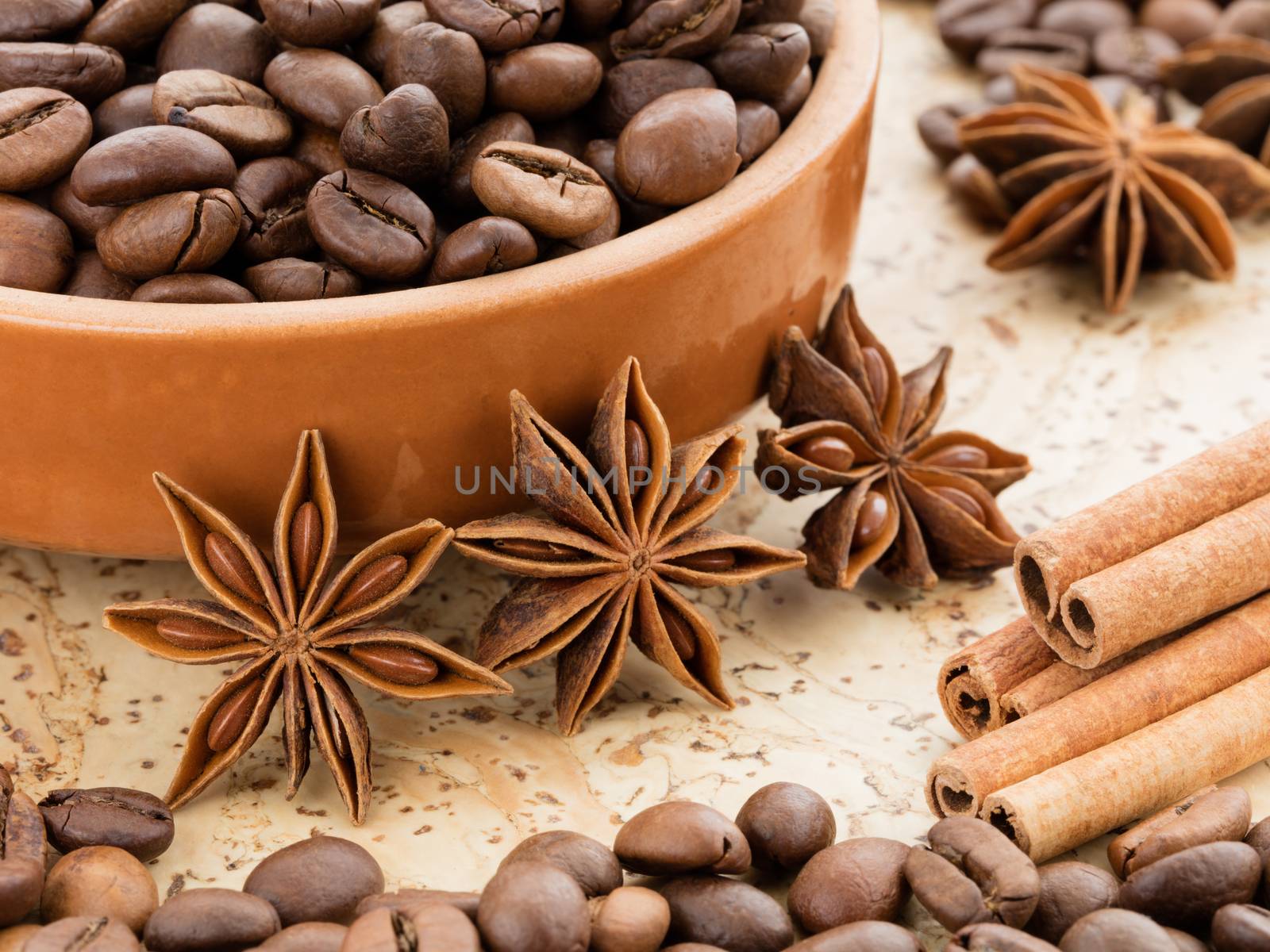 Black coffee beans lying on a clay dish. Cinnamon sticks and star anise star near the saucer of coffee beans on a cork board.