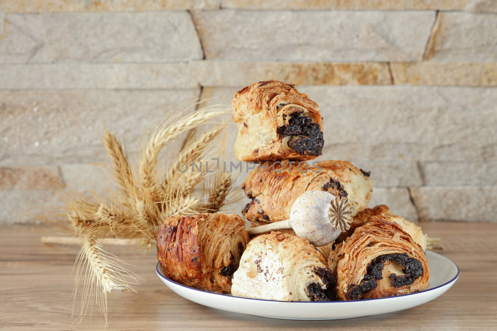 Sweet buns with poppy seeds on a white porcelain plate with a blue rim, on a wooden table and near a stone wall - sandstone. Poppy head and spikelets lie on sweet rolls