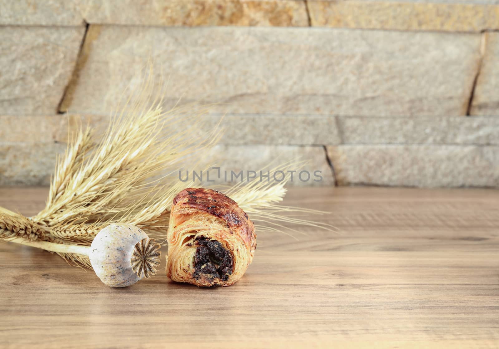 A sweet roll with poppy lies on a wooden table and near a stone wall - sandstone. Poppy head and spikelets lie near sweet roll