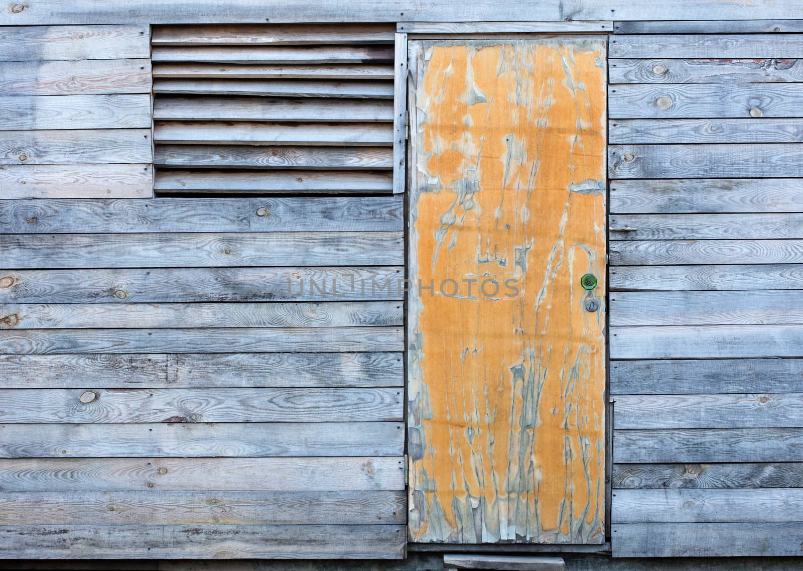 Yellow old door and a window with a wooden grate on the facade of a wooden old gray weather-beaten barn.