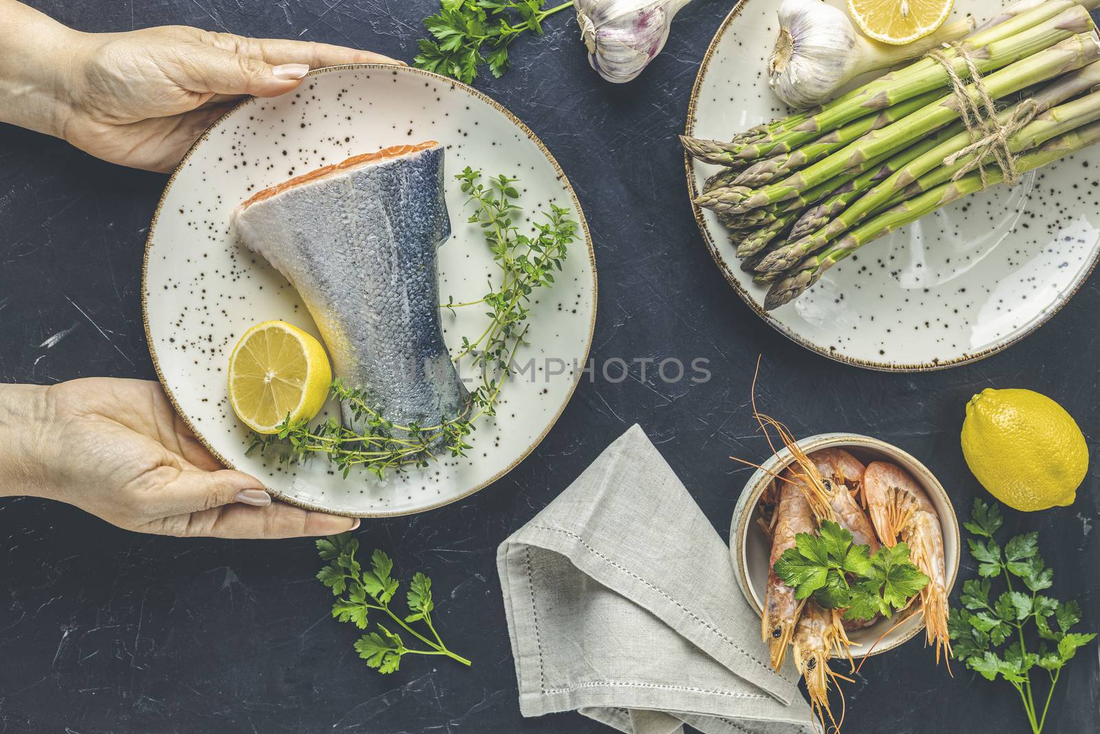 Woman holds ceramic plate with raw trout fish, thyme and lemon in hands on black concrete table surface surrounded plates with fresh raw asparagus, shrimp, prawn, parsley. Healthy seafood background.