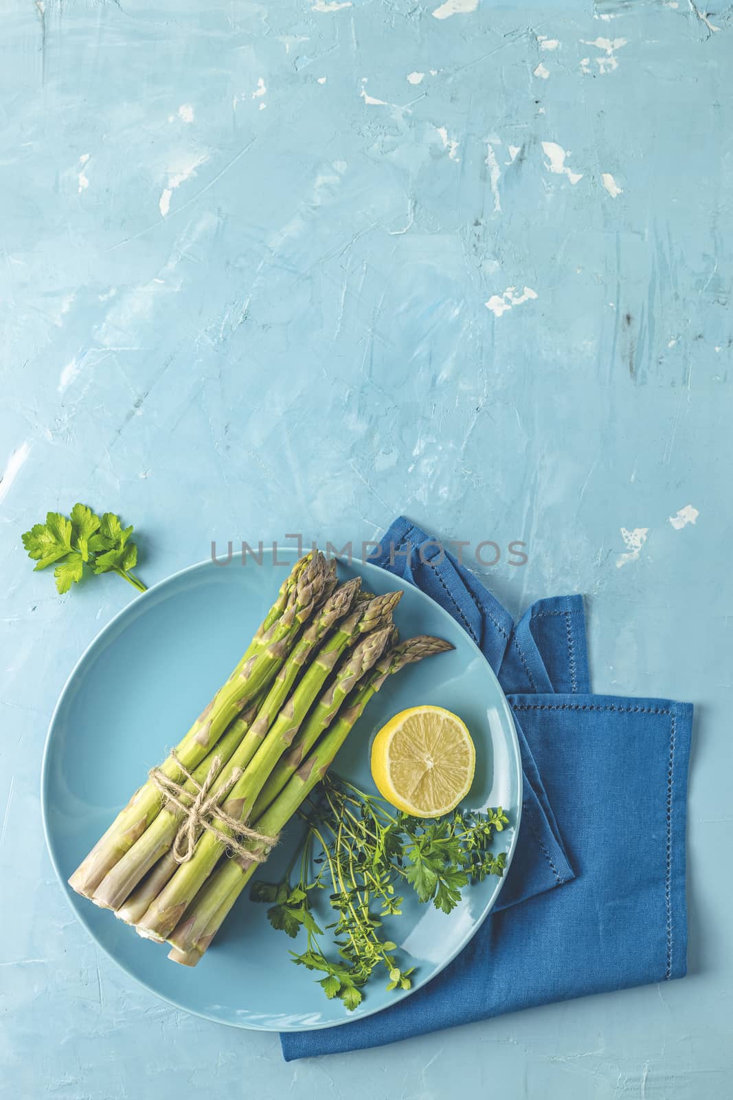 Fresh raw asparagus with lemon and parsley in blue ceramic plate with napkin on light blue concrete table surface. Healthy food background with copy space for you text.