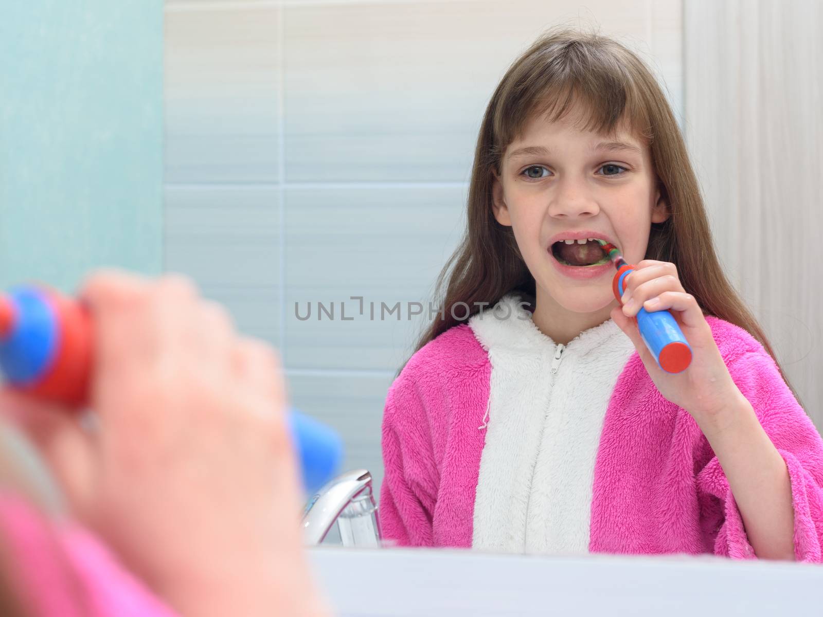 The girl looks in the mirror and brushes her teeth with an electric toothbrush by Madhourse