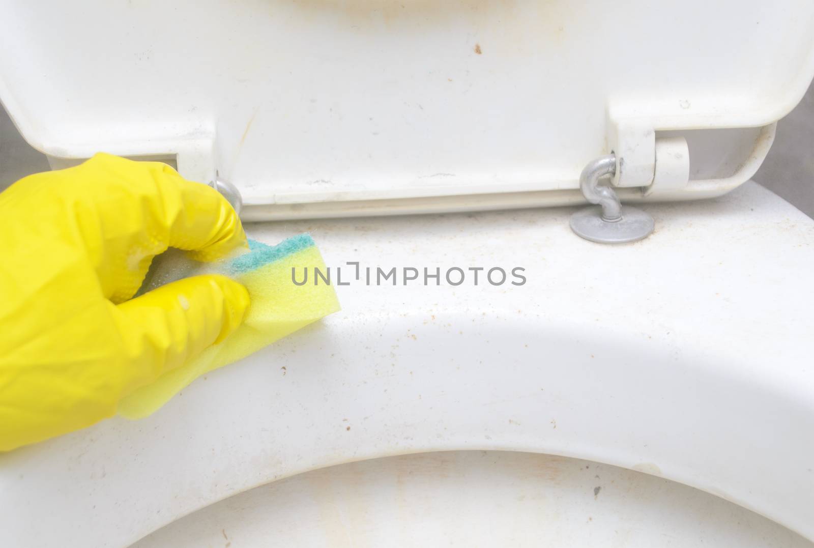 A man in a yellow protective glove washes the dirty rim of the toilet with a sponge in bathroom.Regular domestic cleaning.  by andre_dechapelle