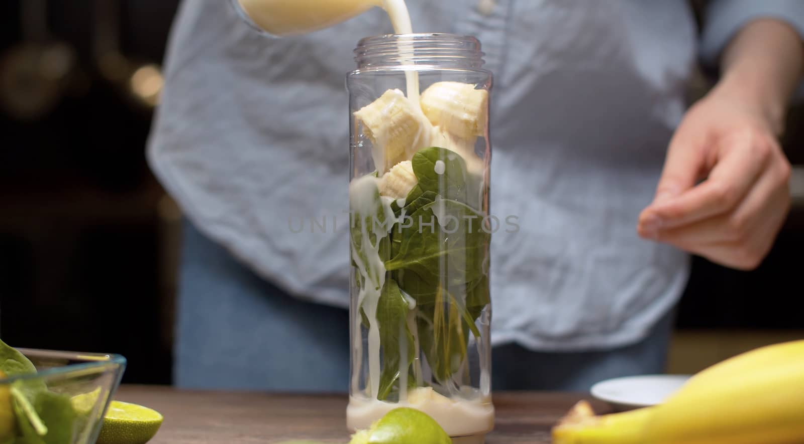 Close up female hands pouring almond milk into the blender glass with banana and spinach leaves. Cooking smoothies in the kitchen. Healthy lifestyle and eating concept