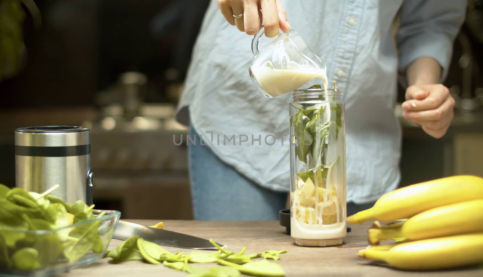 Female hands pouring almond milk in blender glass with banana and spinach leaves. Cooking smoothies in the kitchen. Healthy lifestyle and eating concept