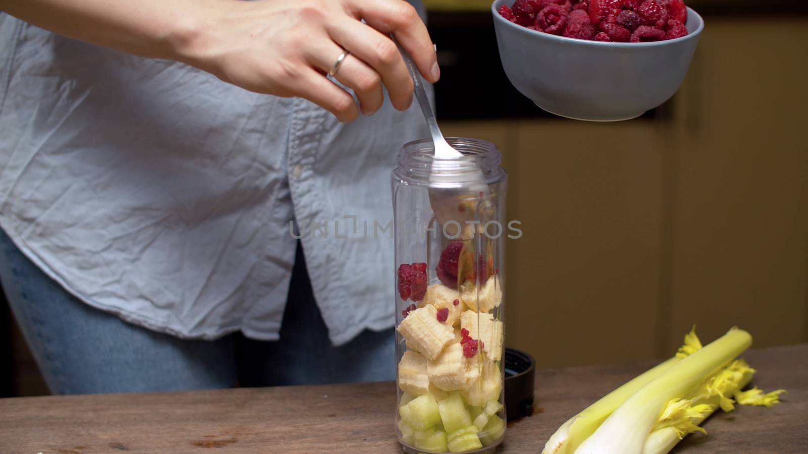 Female hands pouring raspberries in blender glass with banana and celery slices. Healthy lifestyle and eating concept
