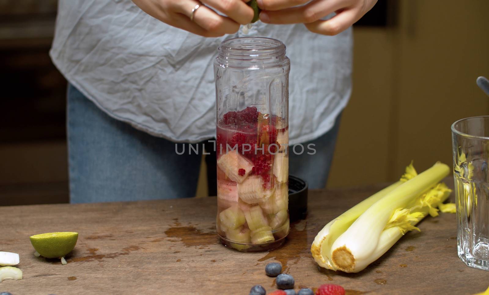 Close up woman's hands squeezing lime juice in blender glass with banana, raspberries and celery slices. Healthy eating concept