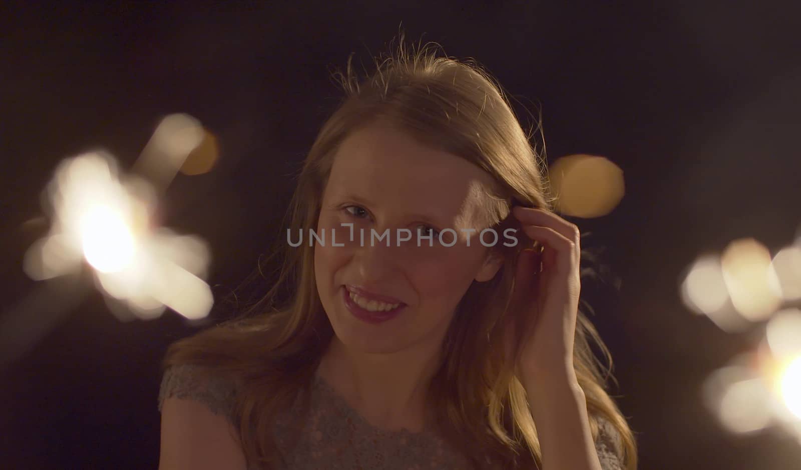 Close up portrait of beautiful caucasian woman celebrating indoors. Blurred sparklers in the foreground. Blond smiling lady on the party looking at camera