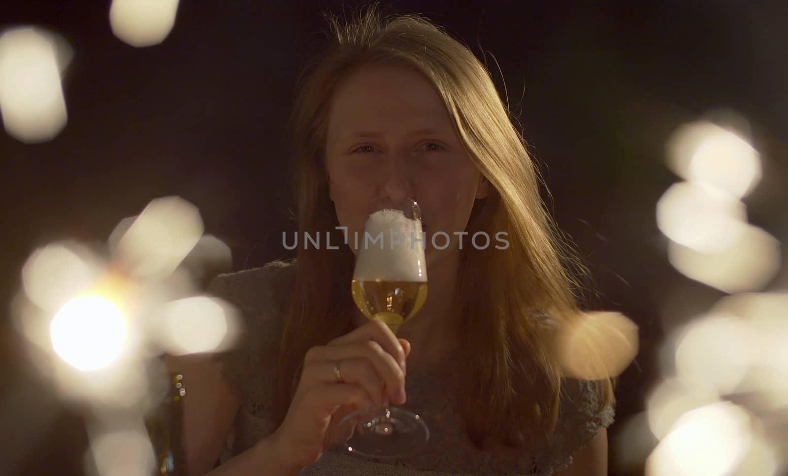 Close up portrait of beautiful young woman drinking champagne on the party. Blurred sparklers in the foreground. Blond smiling woman celebrating indoors. Front view