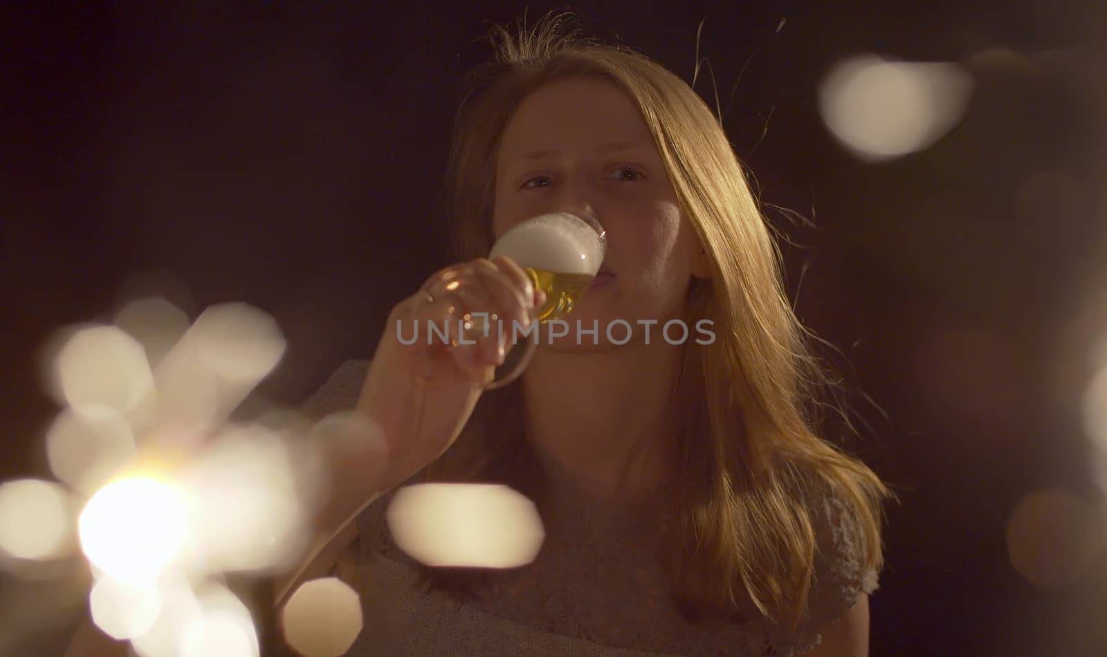 Close up portrait of beautiful young woman drinking champagne on the party. Blurred sparklers in the foreground. Blond smiling woman celebrating indoors. Front view