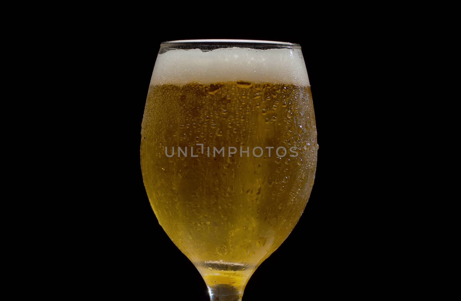 Close up beer in misted transparent glass on black background. Froth on the beer, bubbles beatifully shining in the light ray. Droplets of water on the glass