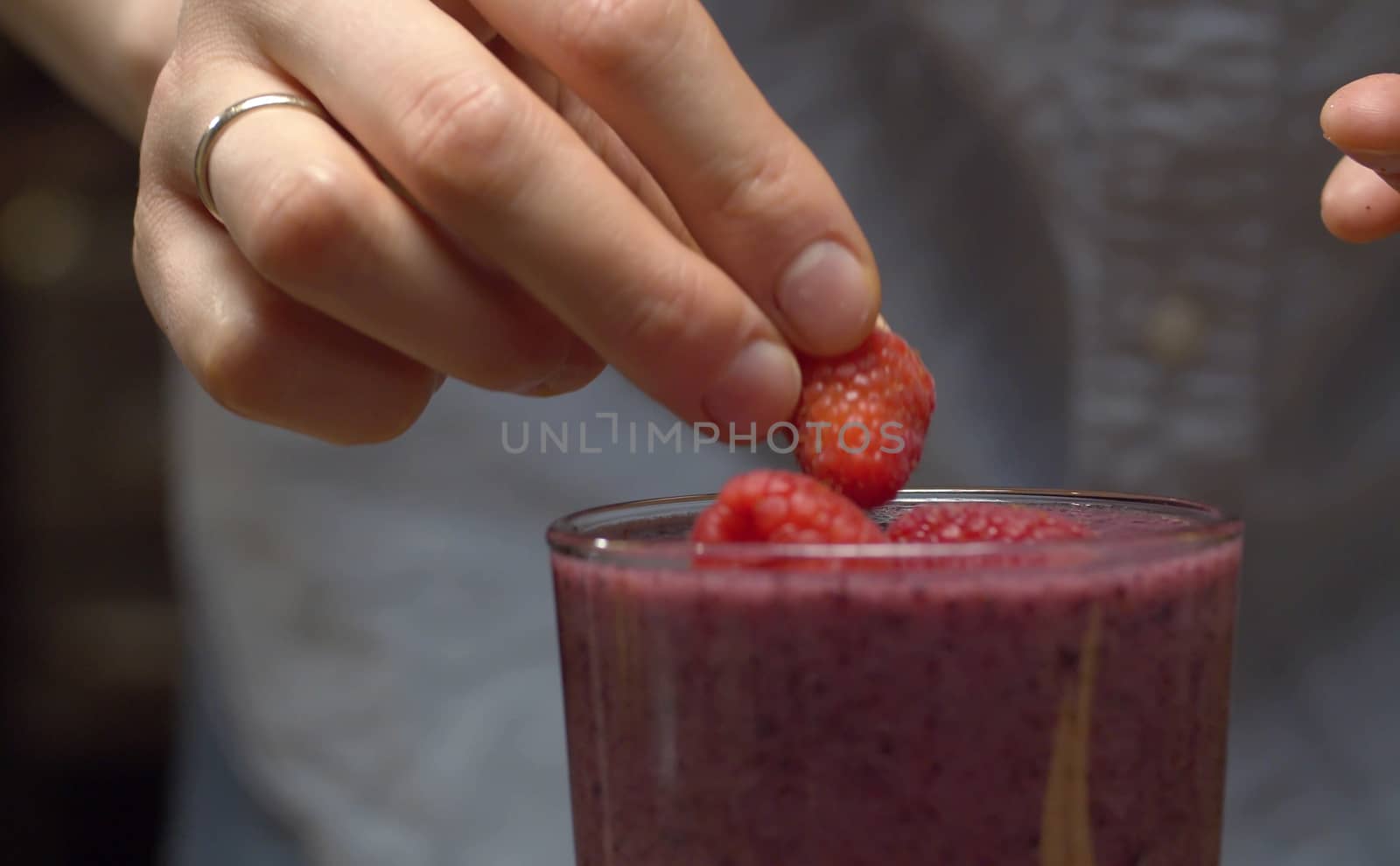 Close up glass of ready made smoothies on the table. Female hands gently decorating it with raspberries and blueberries closeup. Fresh healthy food