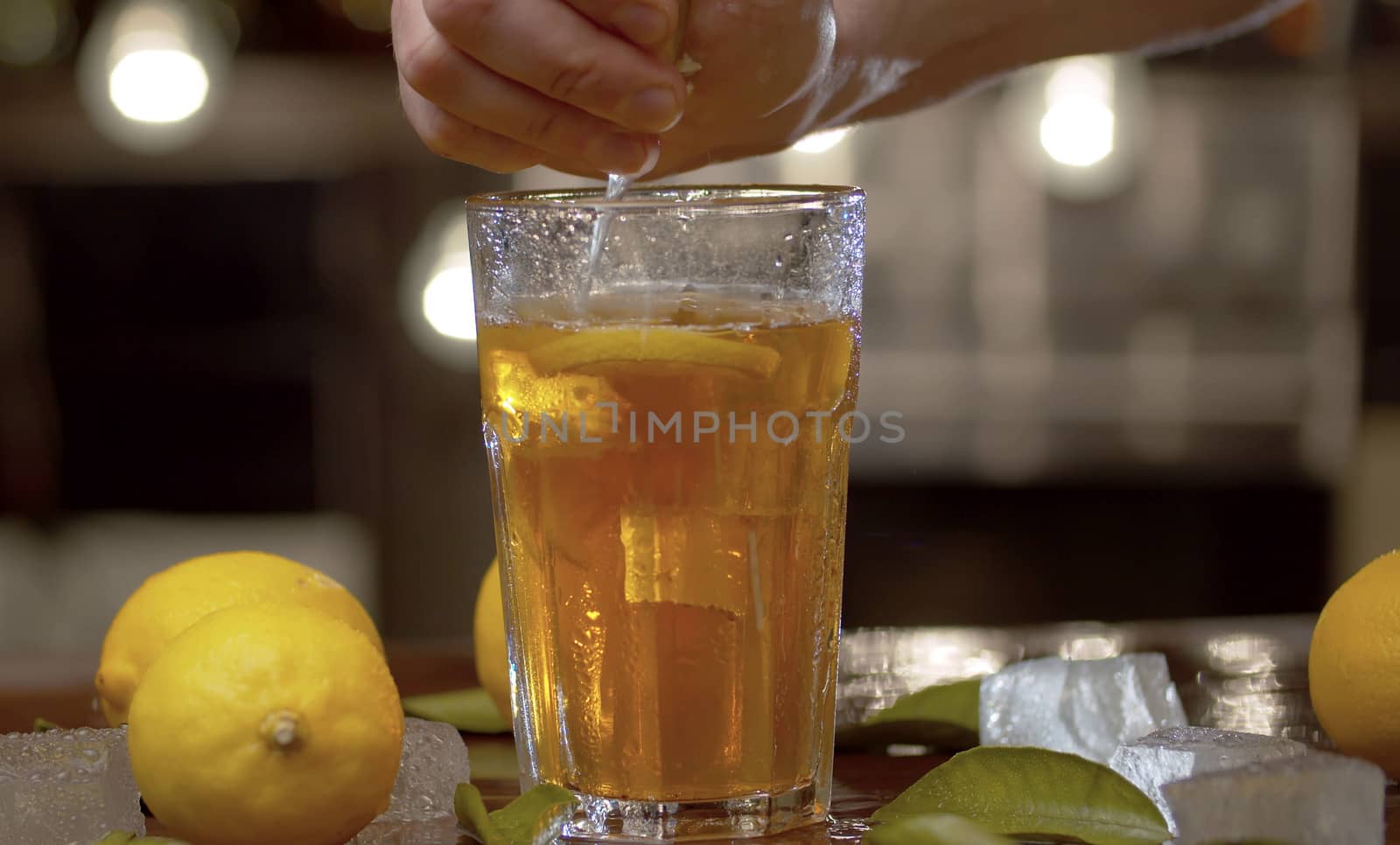 Close up male hand squeezing lemon into a glass with ice tea or cocktail. Glass with a refreshing drink, ice, lemons and leaves on a table against the blurry lights background