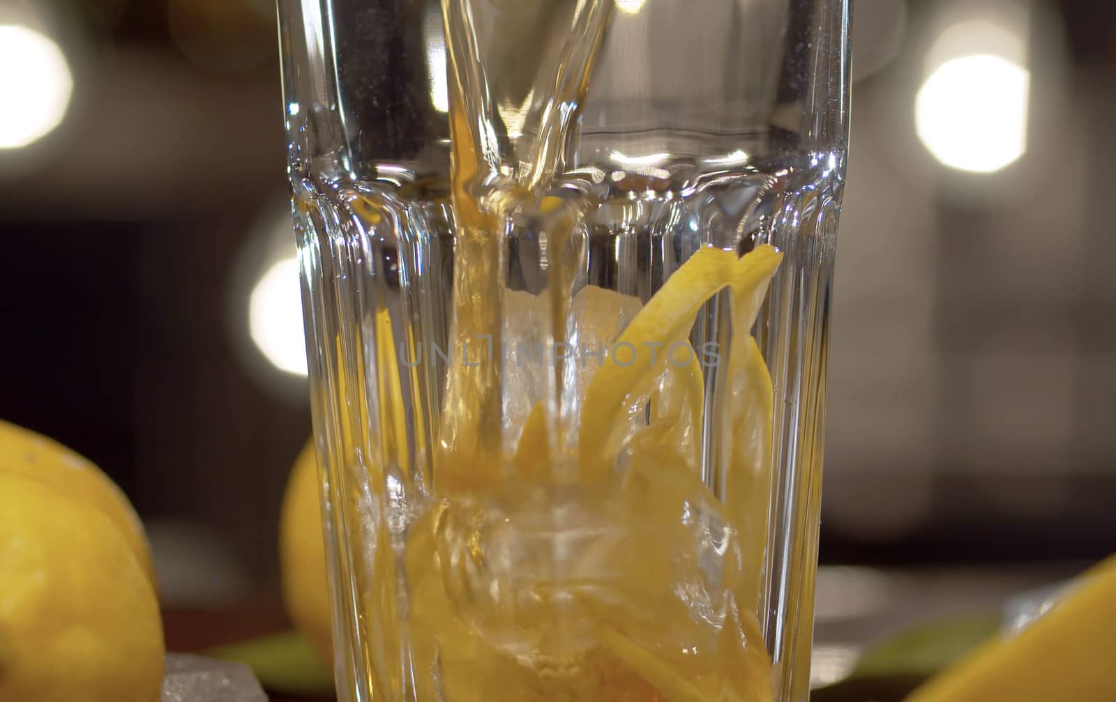 Close up tea pouring into the glass with ice and lemon. Glass with ice tea or cocktail, ice, lemons and leaves on a table against the blurry lights background. Bar counter closeup