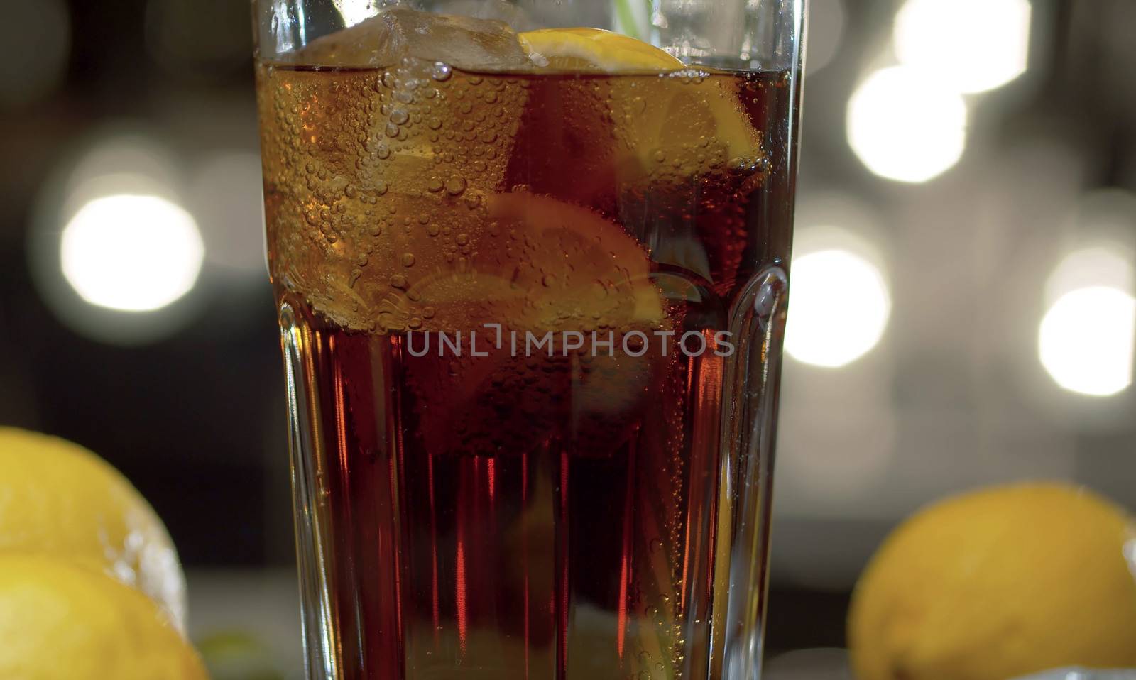 Close up glass on the table. Glass with cocktail or fizzy sweet drink with ice and lemon on a table against the blurry lights background.