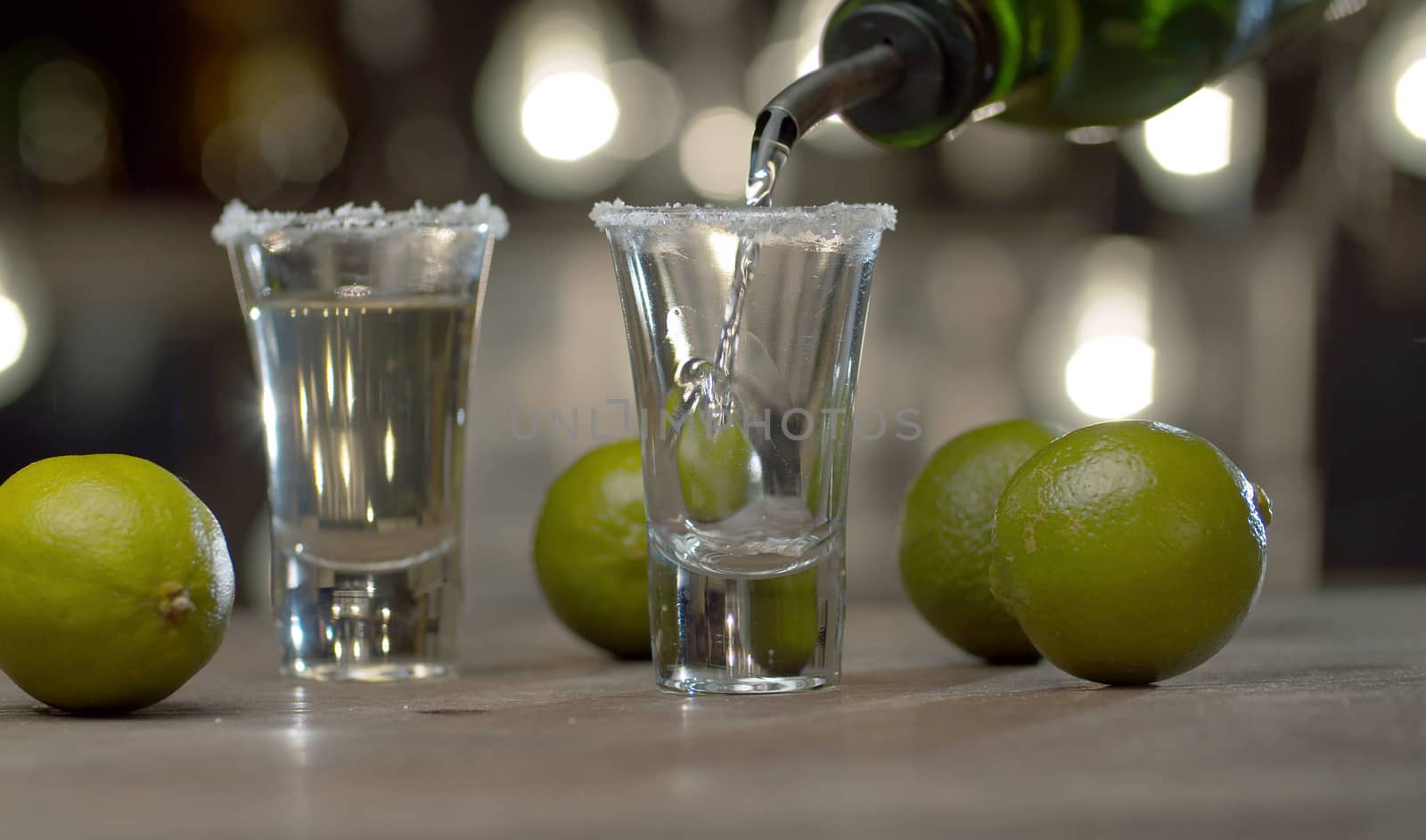 Close up bartender pouring tequila from the bottle into the glasses. Bar counter on the background of blurry light bulbs.