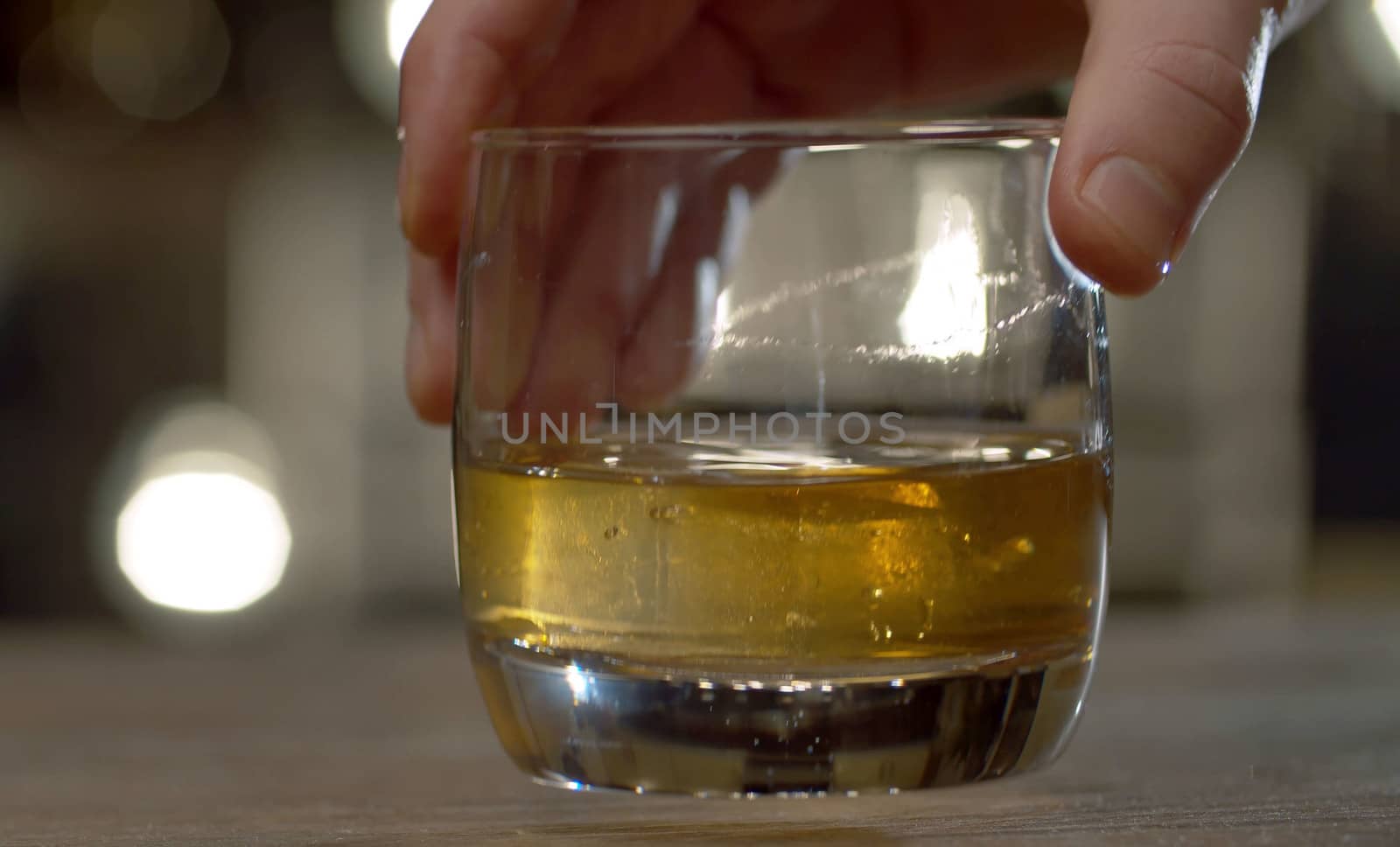 Close up bartender's hand stirring whiskey in drinking glass with ice cubes. Bar counter on the background of blurry light bulbs.