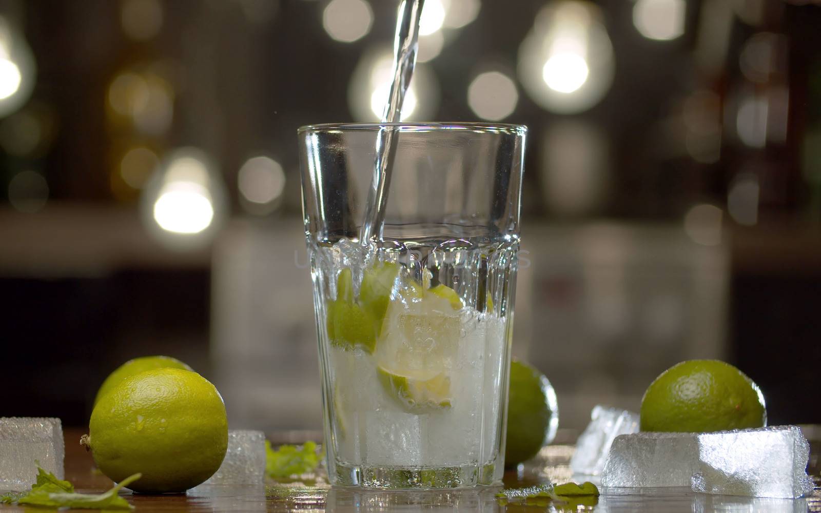 Close up fizzy water pouring into a glass with lime and ice on a blurry lights background. Tonic, soda or mineral water.