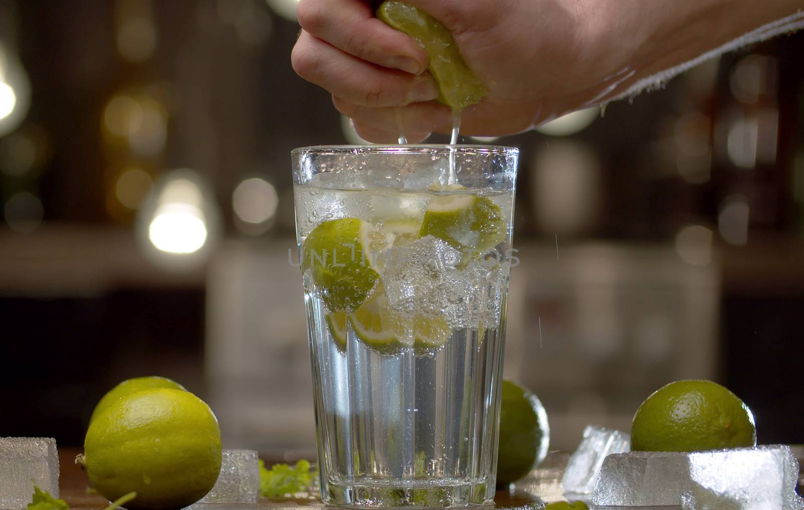 Close up male hand squeezing lime into a glass with tonic, soda water or cocktail. Glass with a refreshing drink, ice, limes and leaves on a table against the blurry lights background
