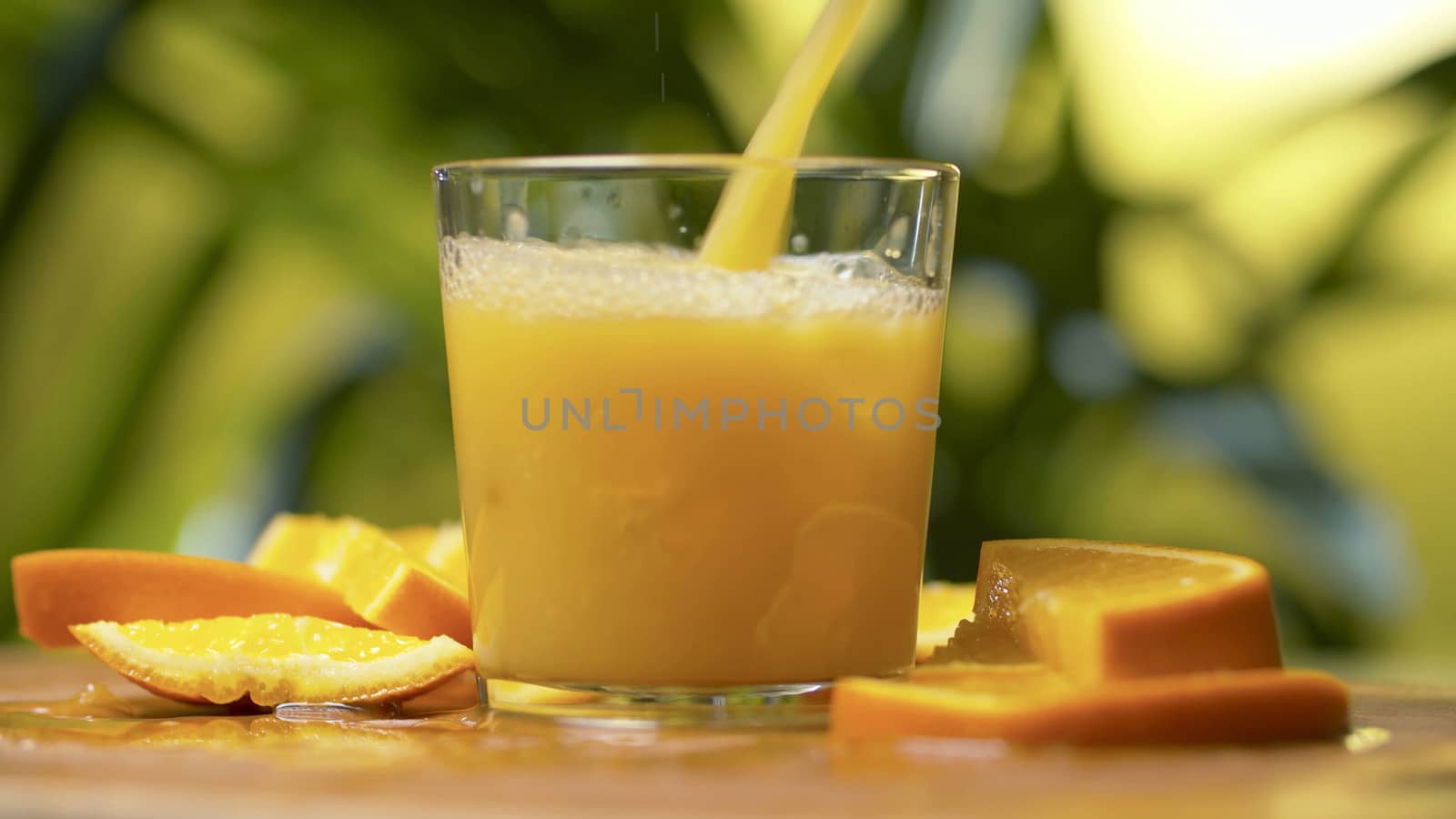 Fresh orange juice pouring into a transparent glass. Close up refreshing yellow beverage on natural background. Sliced oranges on a table. Healthy lifestyle concept.
