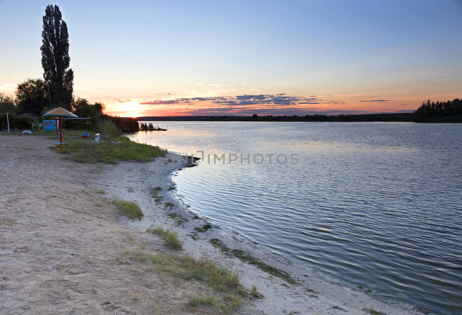 On a river beach, a summer bright sunset and a reflection of the sun's rays on the calm water of the river by Sergii