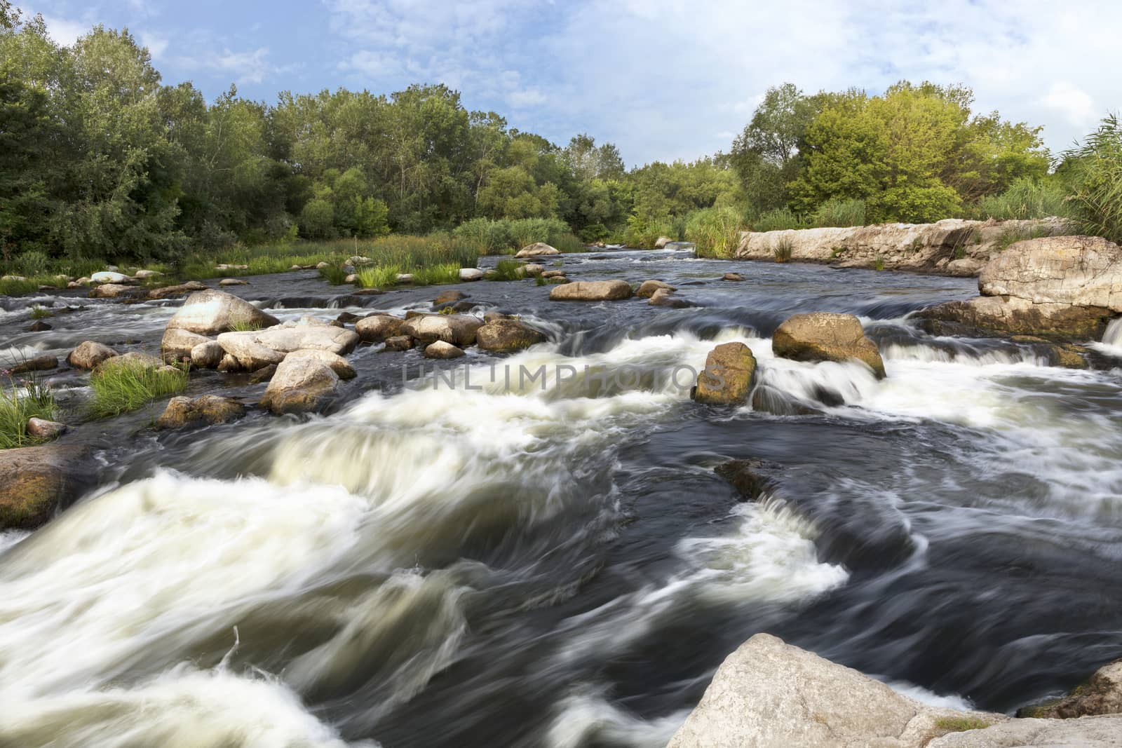 The rapid flow of the river, rocky shores, rapids, bright green vegetation and a cloudy blue sky in the summer. Ukraine