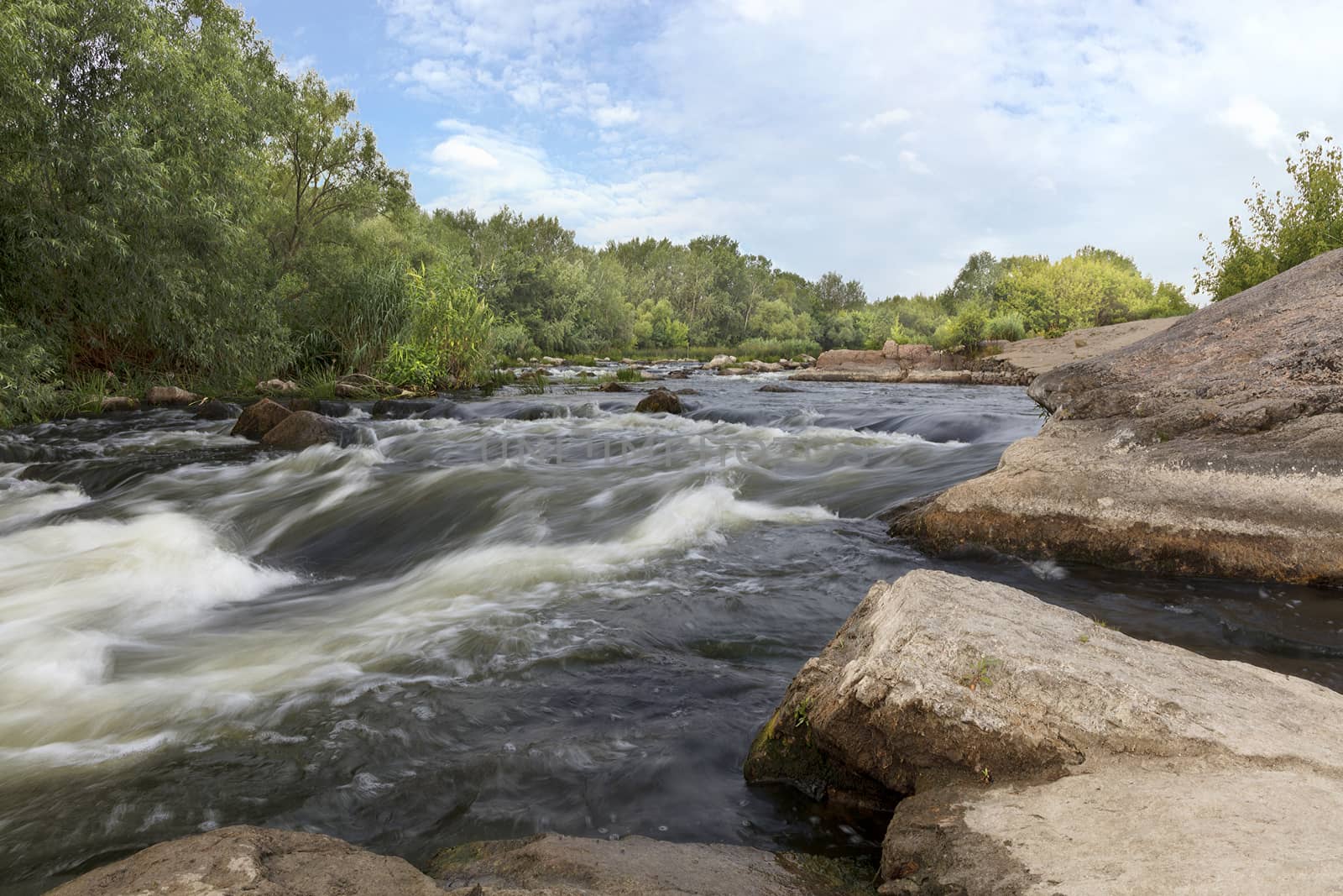 Rocky shores, rapids, fast river flow, bright green vegetation and a cloudy blue sky in summer