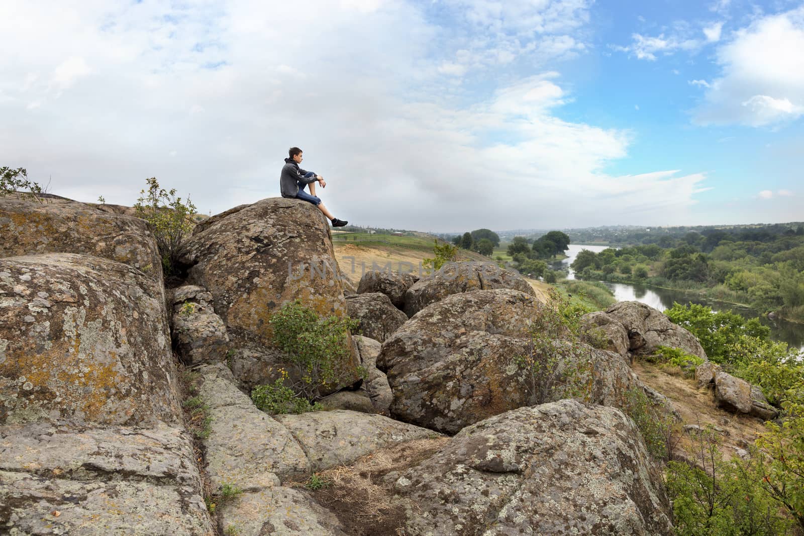 The teenager sits on top of a large stone boulder on the bank of the South Bug River and looks at the river below. The river Southern Bug in the summer - river flow, rocky shores, bright green vegetation and a cloudy blue sky