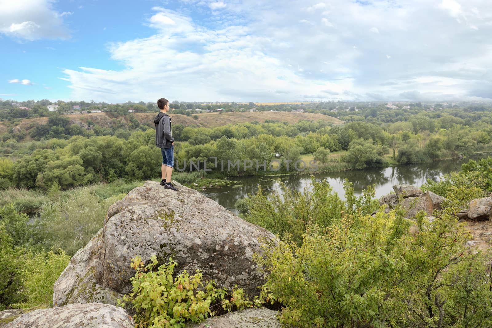 The teenager stands on top of a large stone boulder on the bank of the South Bug River and looks at the river below. The river Southern Bug in the summer - river flow, rocky shores, bright green vegetation and a cloudy blue sky