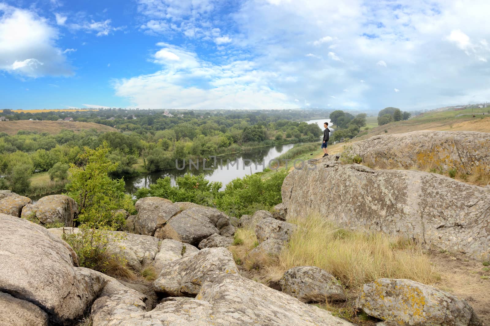 The teenager stands on top of a large stone boulder on the bank of the South Bug River and looks at the river below. The river Southern Bug in the summer - river flow, rocky shores, bright green vegetation and a cloudy blue sky