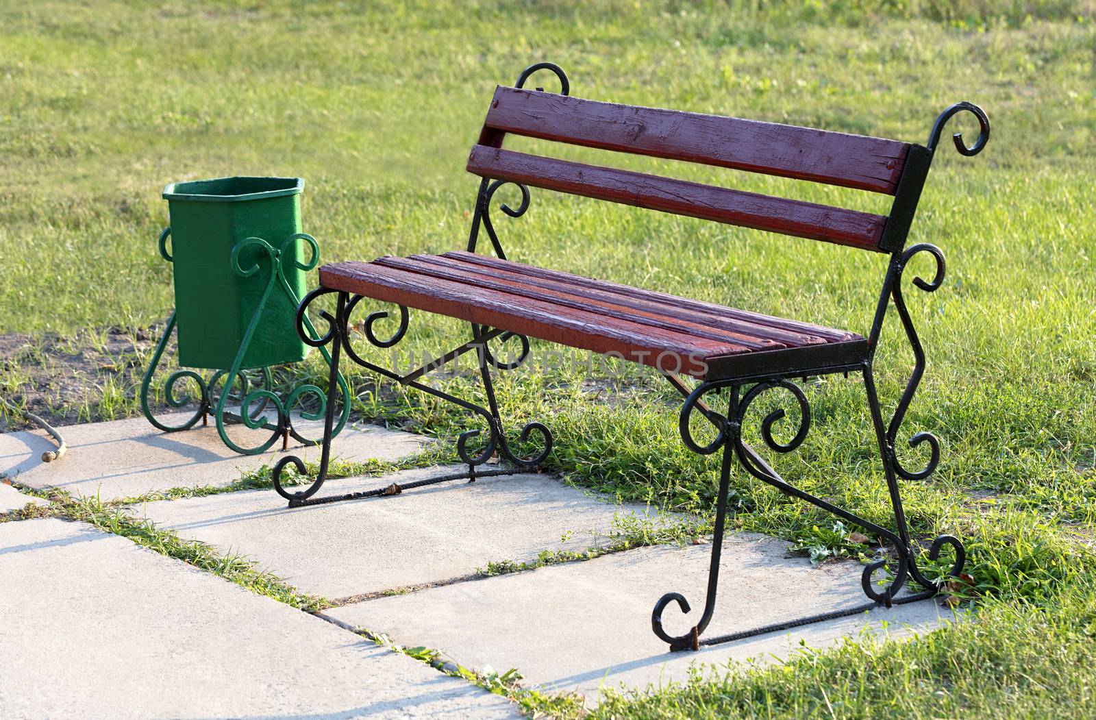 A wooden bench and a garbage can in antique style stand on the green lawn of the public park.