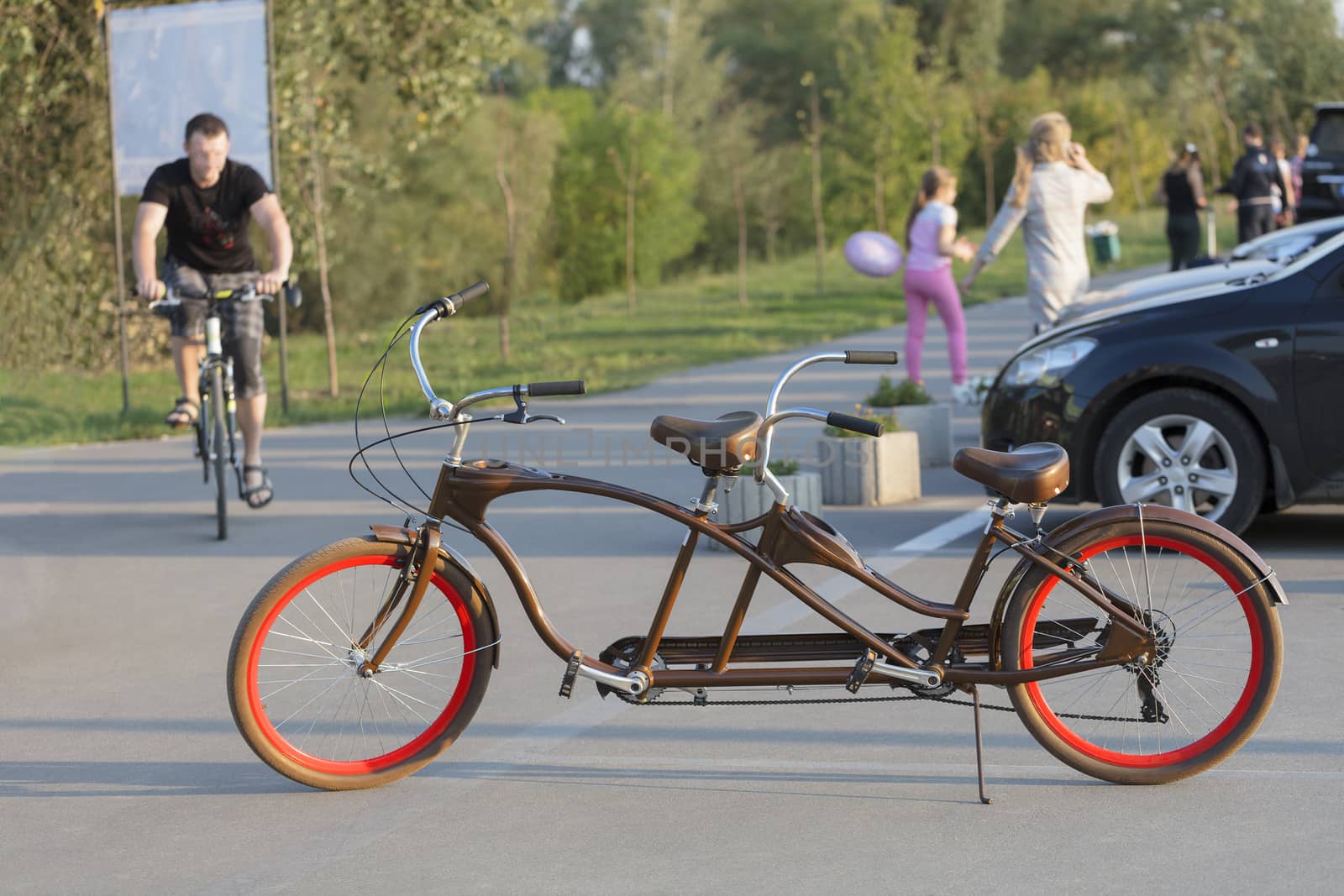 A chocolate-colored tandem bicycle with scarlet wheels is parked in a car park near the bike path in the evening sun.