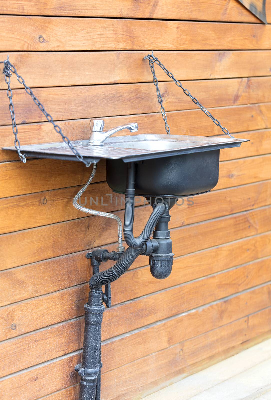 A modern washbasin sink with a stainless steel faucet installed outside on a chain on the wall of a wooden house, in a rustic farmhouse