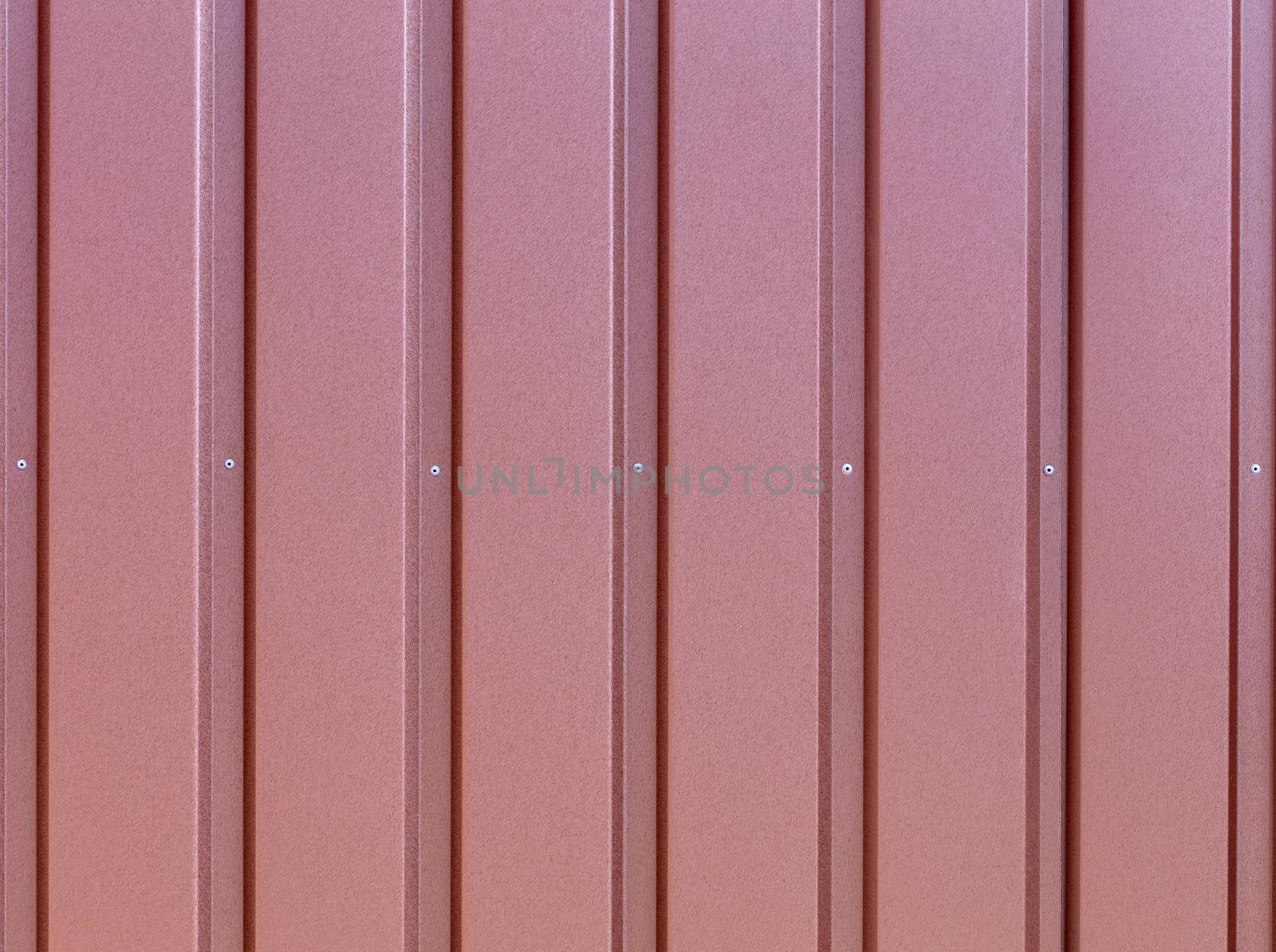 Reddish-brown metal fence made of corrugated steel sheet with vertical guides and metal rivets in the middle. Corrugated reddish-brown iron sheet background close up.