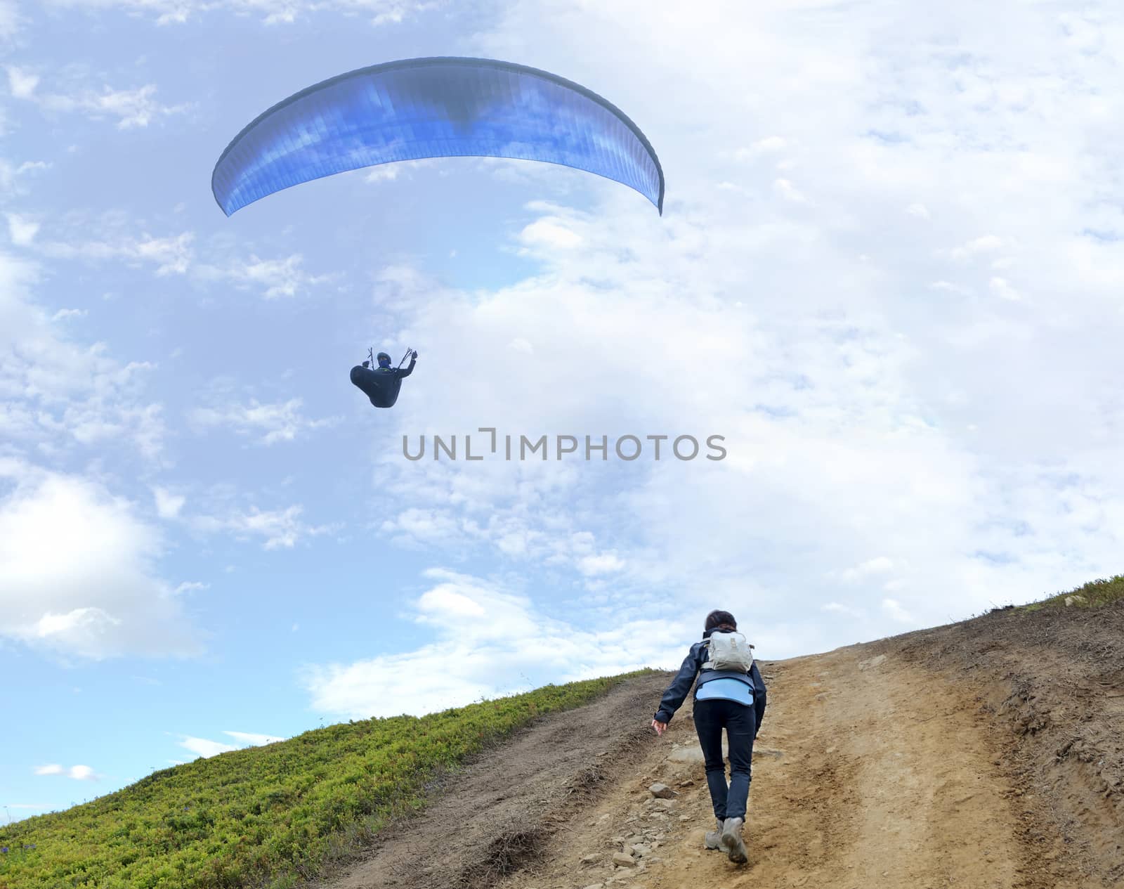 A young woman with a backpack behind her shoulders climbs up a mountain and looks at the floating paraglider hovering in the air against the blue sky and white clouds