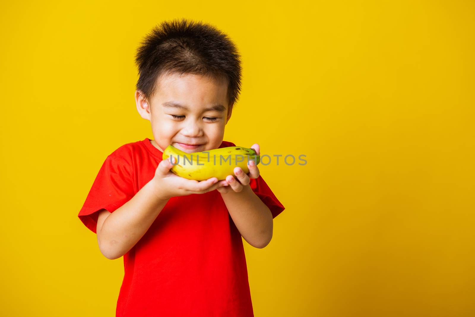 Happy portrait Asian child or kid cute little boy attractive smile wearing red t-shirt playing holds banana fruit, studio shot isolated on yellow background